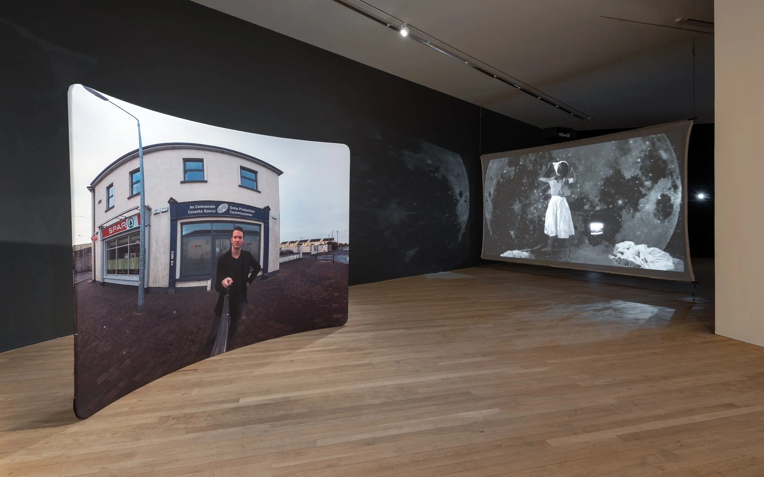 Art installation with two hanging screens, one showing a color photo of a man standing in front of a building, the other displaying a black-and-white image of a woman with the moon in the background.