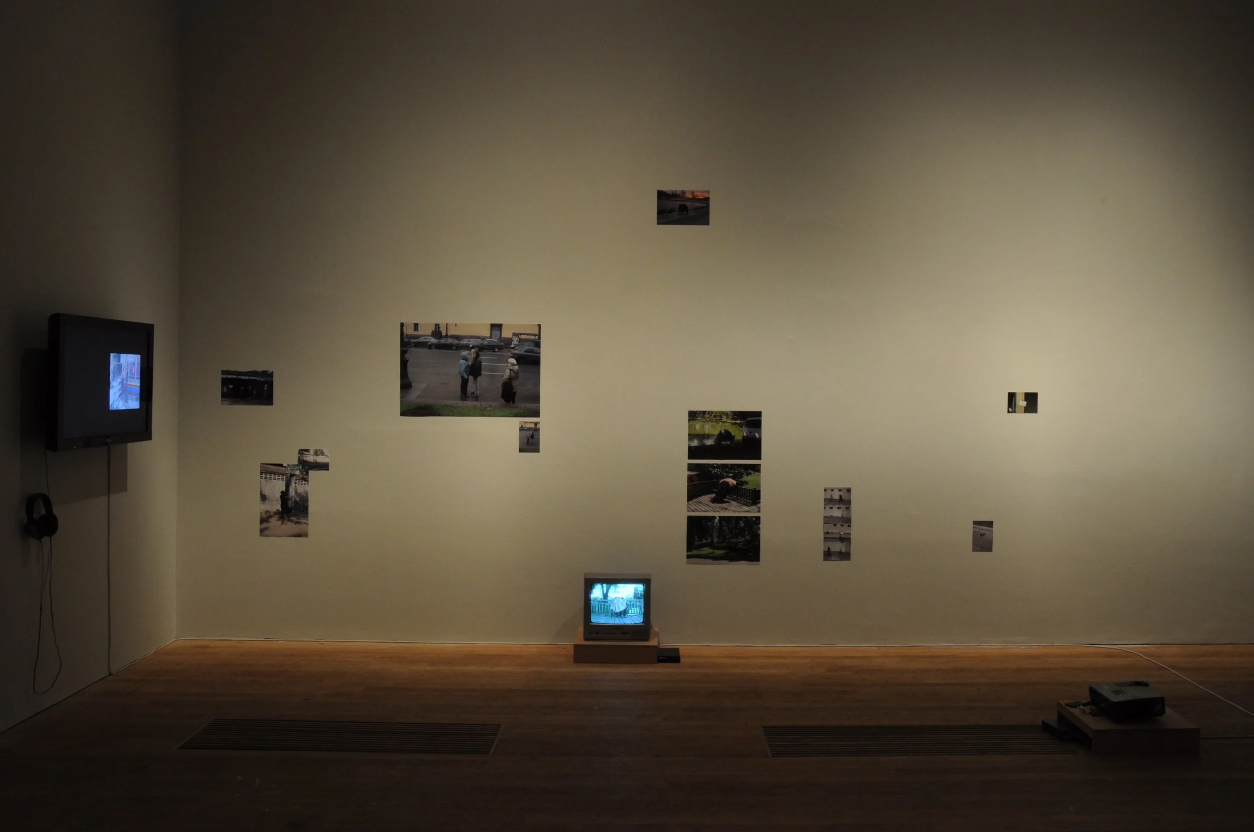 An art installation in a gallery featuring photographs mounted on a white wall, with a small television on a stand below and a larger television on the left wall.