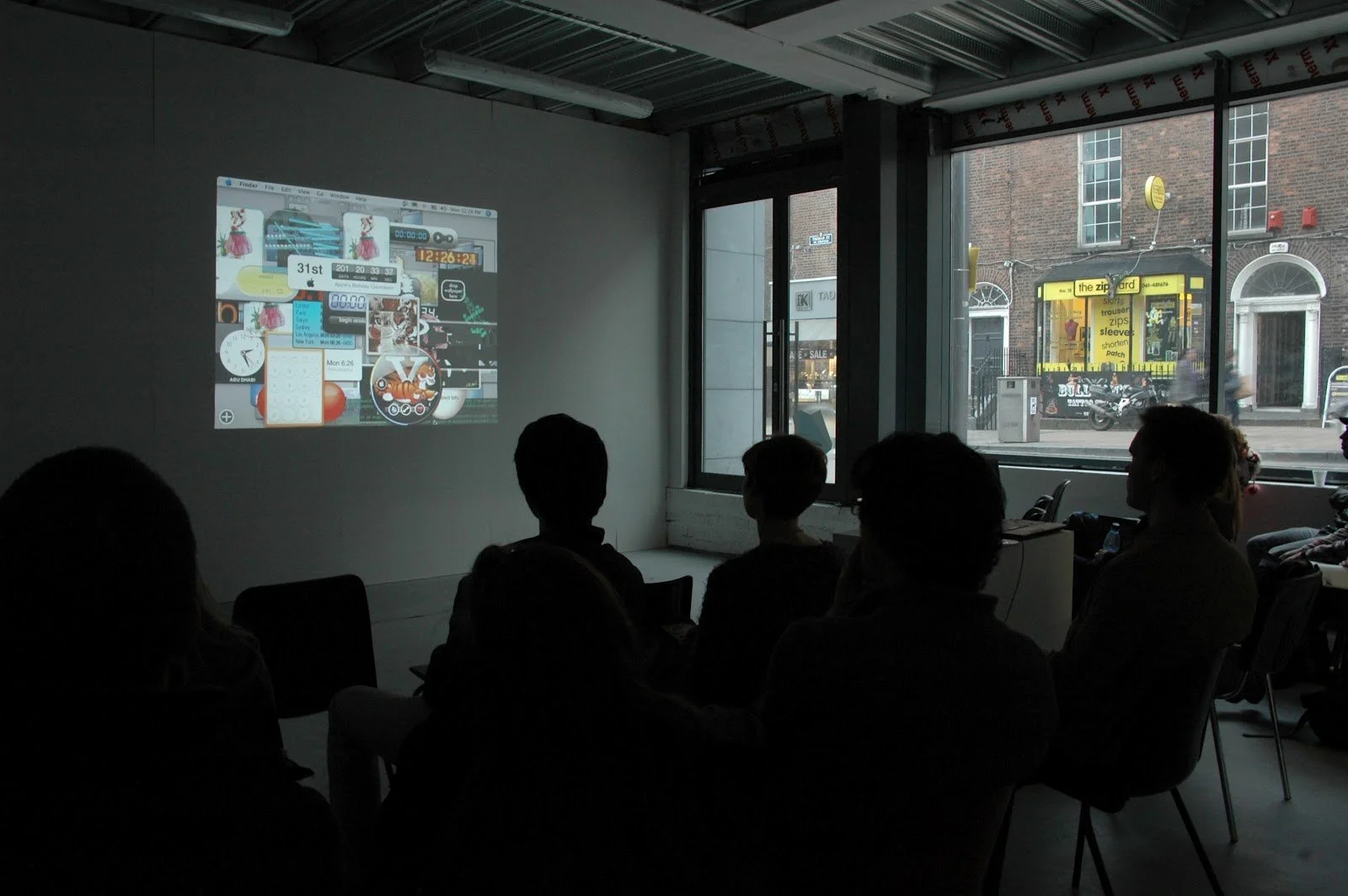 A group of people sitting in a dark room watching a presentation or video projected on a wall. Outside, a city street with stores and pedestrians is visible through large windows.