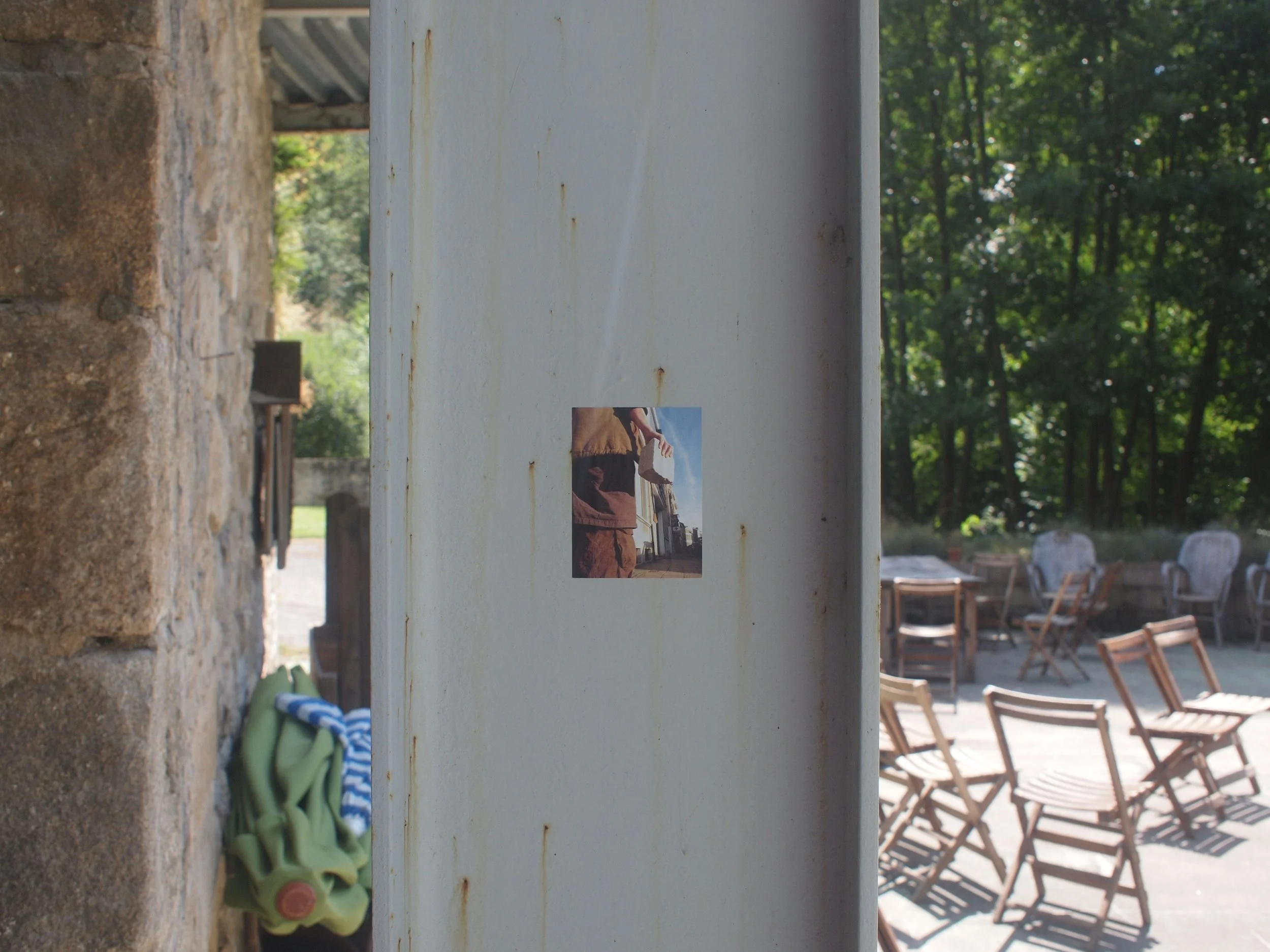 Photo of an outdoor patio area with chairs and tables, partially obscured by a central white wall with a small photo sticker. The background features green trees and a stone wall on the left.