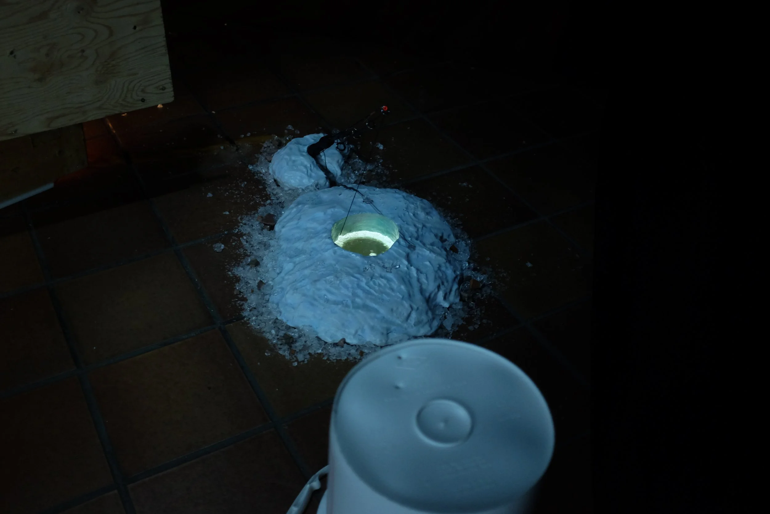 A hole in the tiled floor with melted ice and water spilled around it, a speaker cone with a greenish glow inside, and a white air purifier in the foreground.