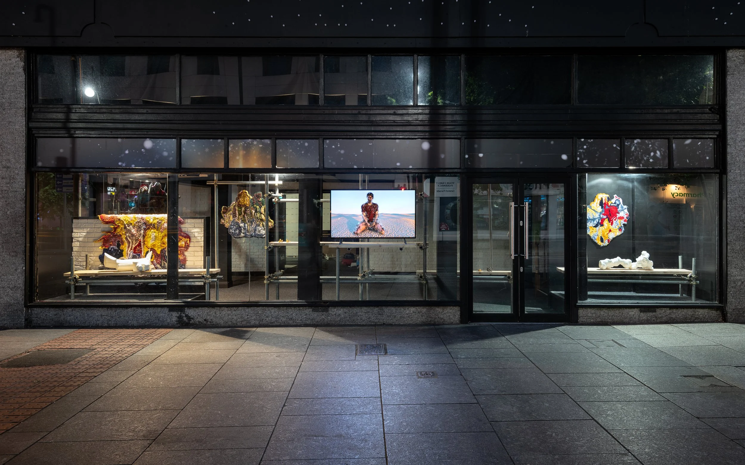 View of an art gallery storefront at night with large glass windows displaying colorful abstract sculptures and a digital screen showing a person sitting in a landscape.
