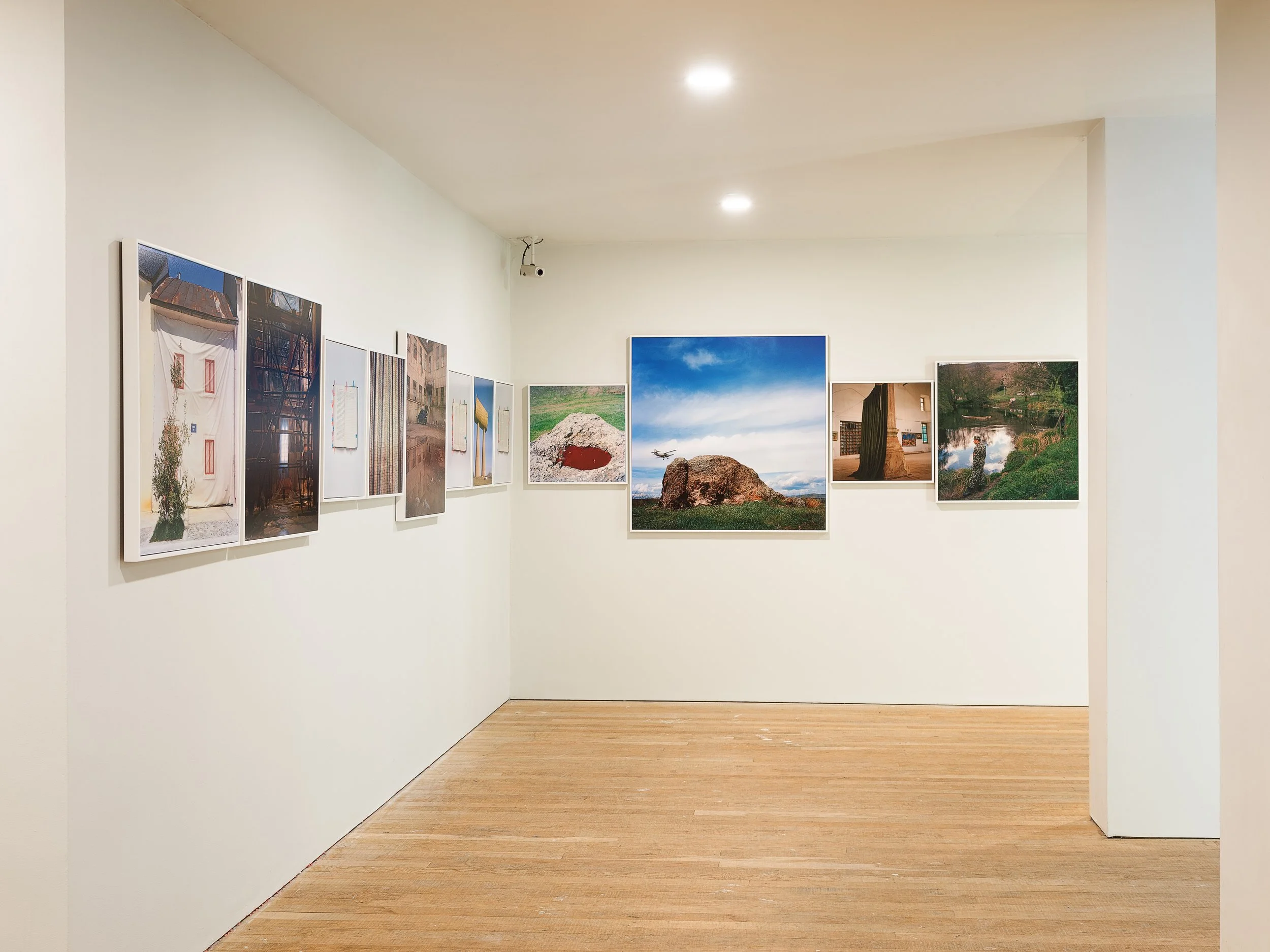 Art gallery with framed photographs and paintings on white walls, wooden floor, and ceiling lights.