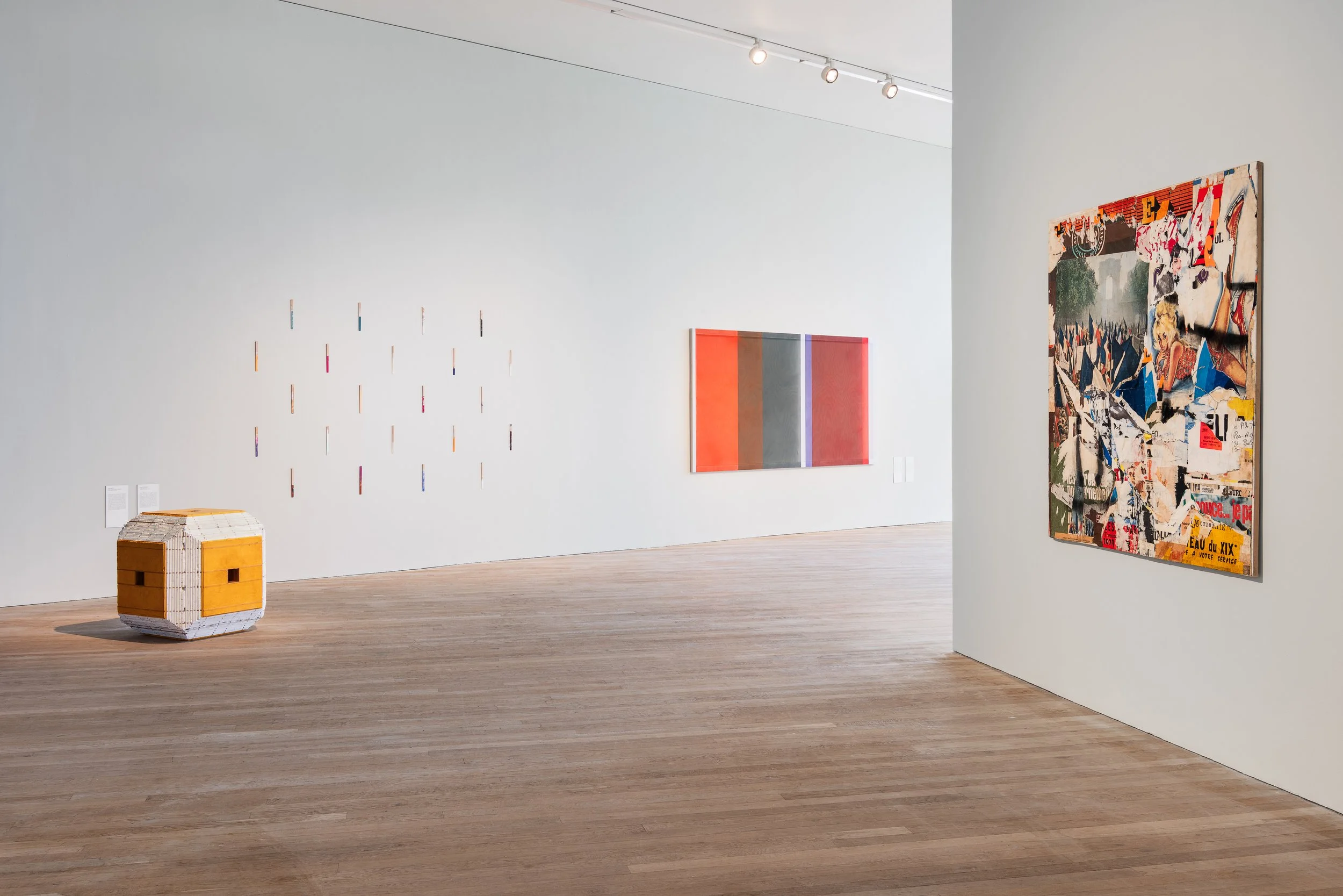 Art gallery with white walls and wooden floors, displaying three modern art pieces: a small sculpture resembling a cube on the floor, a colorful grid of small vertical elements on the wall, and two large abstract paintings with bright colors on the w
