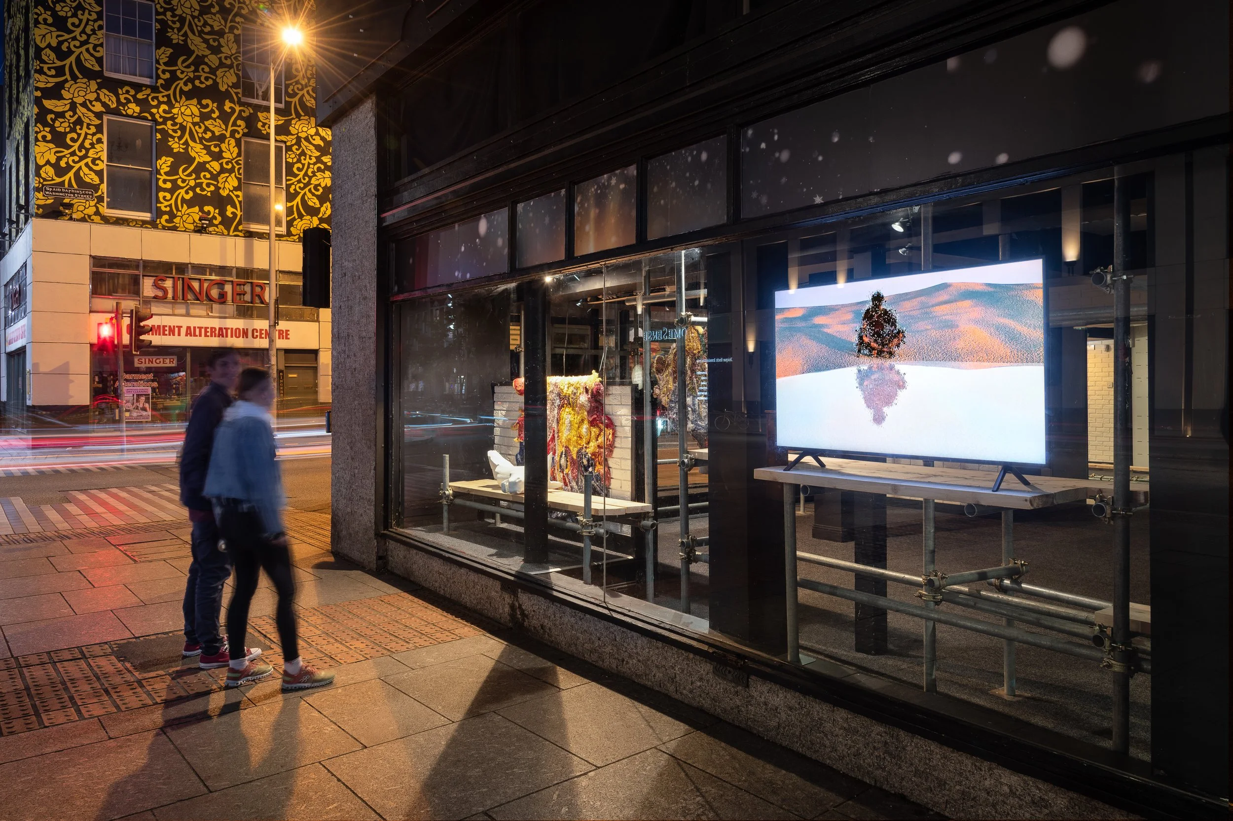 Two people walking past a storefront window at night, with a large digital screen displaying a snowy landscape inside the store, with outside traffic and illuminated signs visible.
