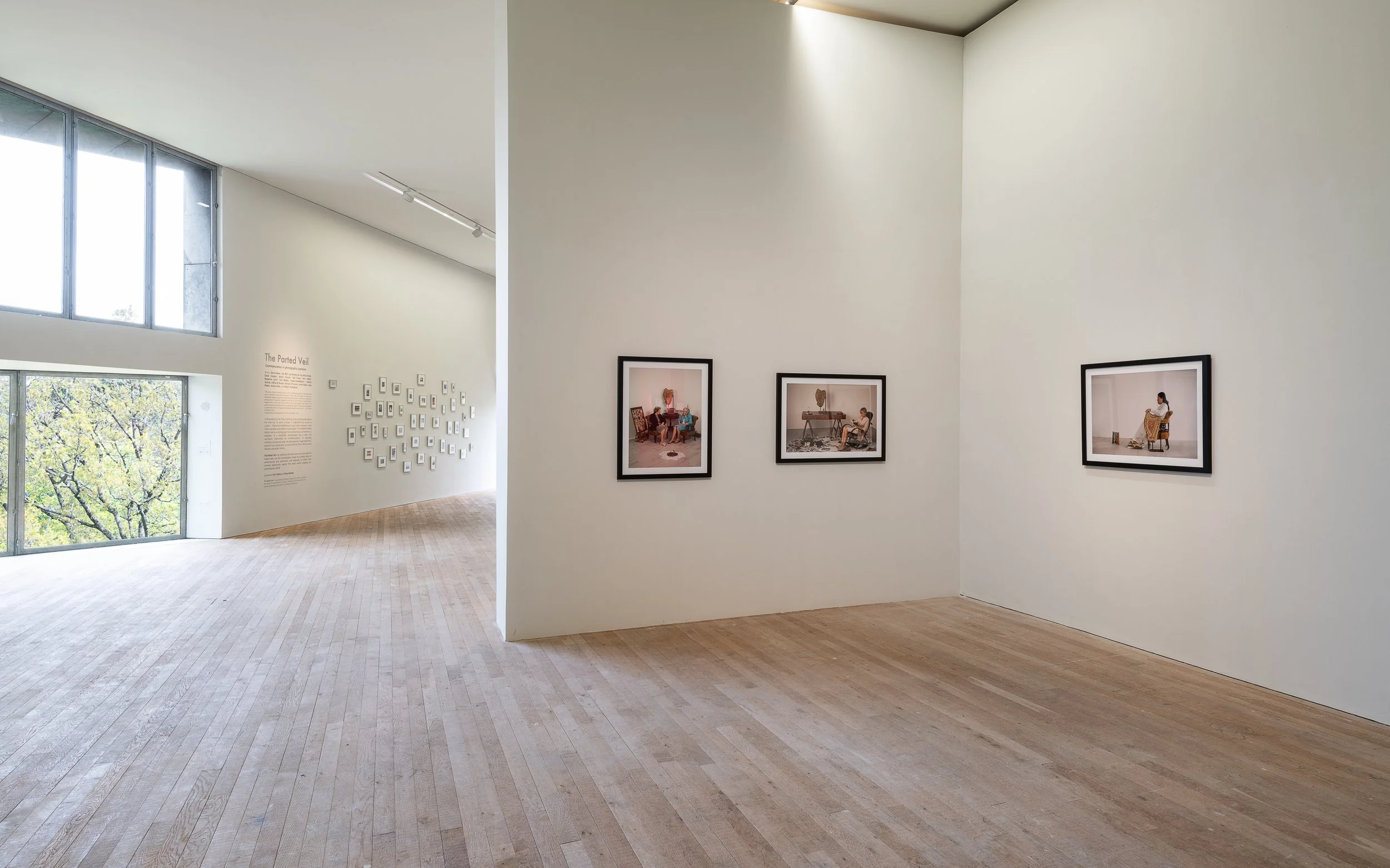Empty art gallery with framed photographs on the walls and a display of small framed artworks on the left wall, large windows letting in natural light, and light wooden floors.