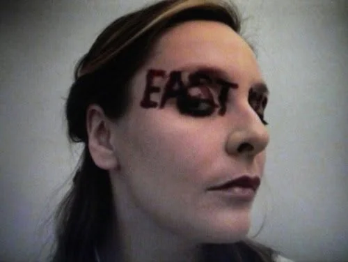 A woman with the word 'EAST' written across her forehead in dark ink, looking to the side with a neutral expression.
