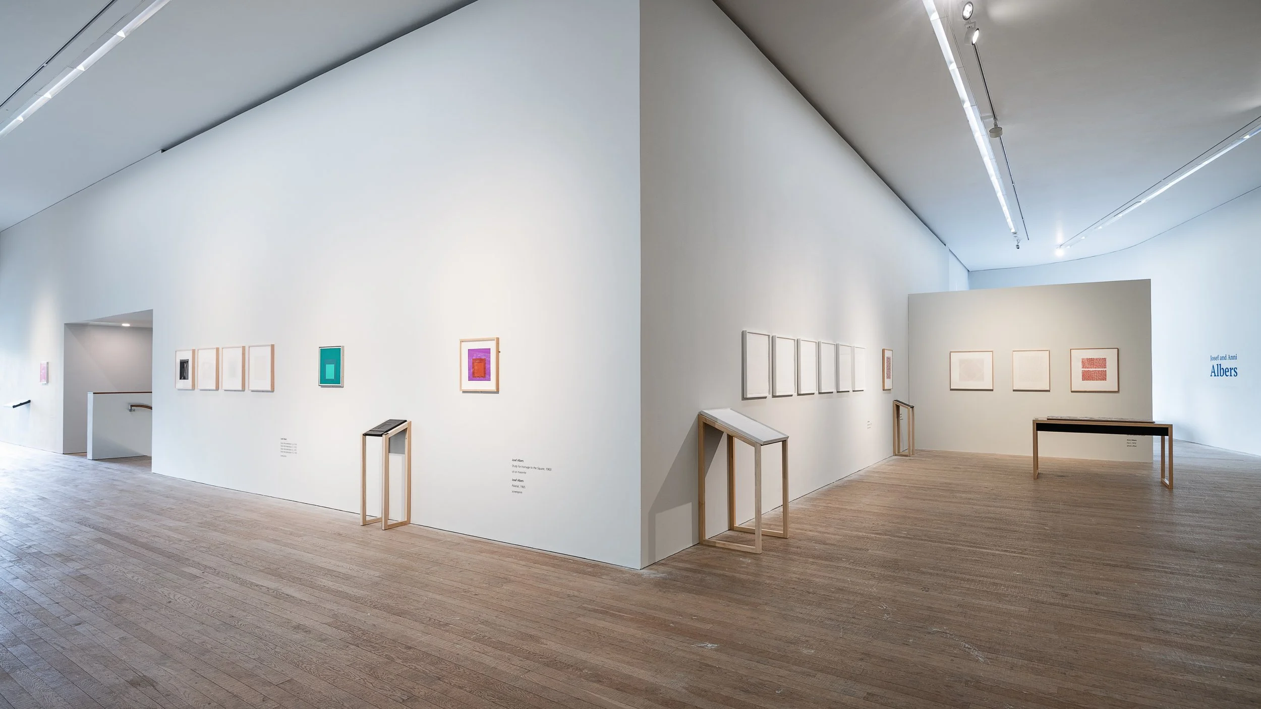 An art gallery with white walls, wooden flooring, and multiple framed artworks. The paintings are abstract, colorful, and minimalist, arranged in a row with some displayed on small wooden stands underneath. Lighting fixtures highlight the artworks, a