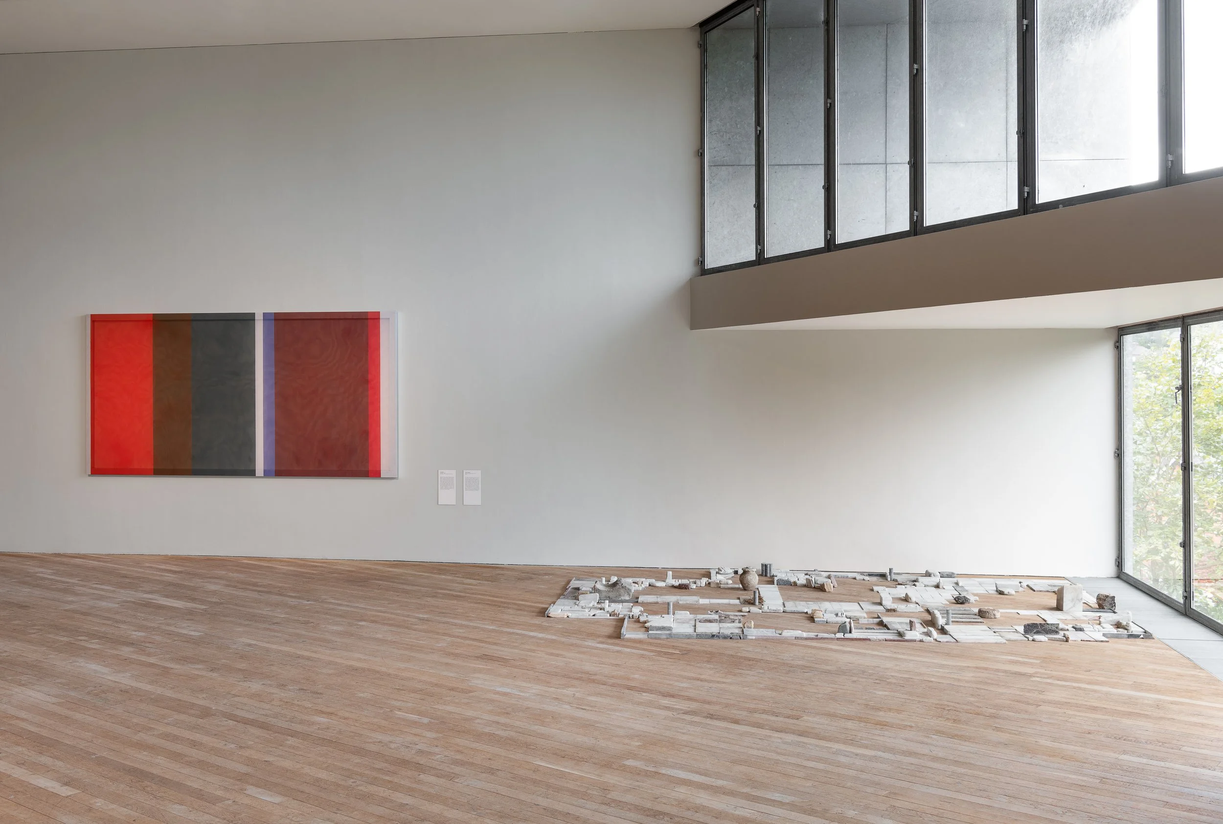 Modern art gallery with a large abstract painting on the wall, a wooden floor, and an installation of white bricks on the floor near a large window.