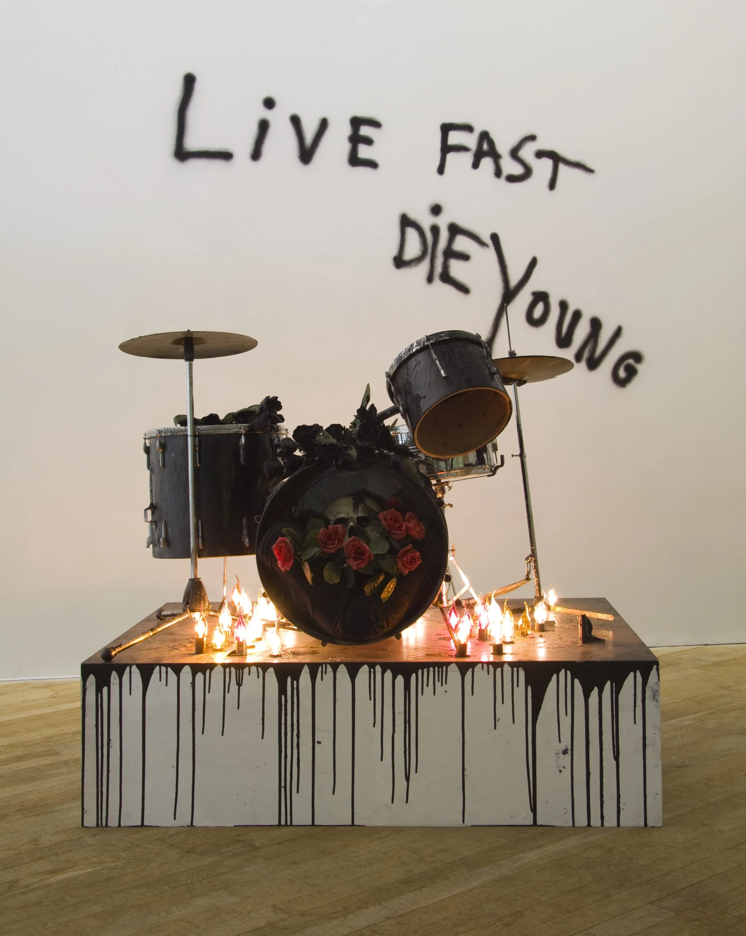 A sculpture of a drum set with candles and roses on the bass drum, placed on a platform with black dripping paint design. Behind it, words 'Live Fast Die Young' are written in black on the wall.