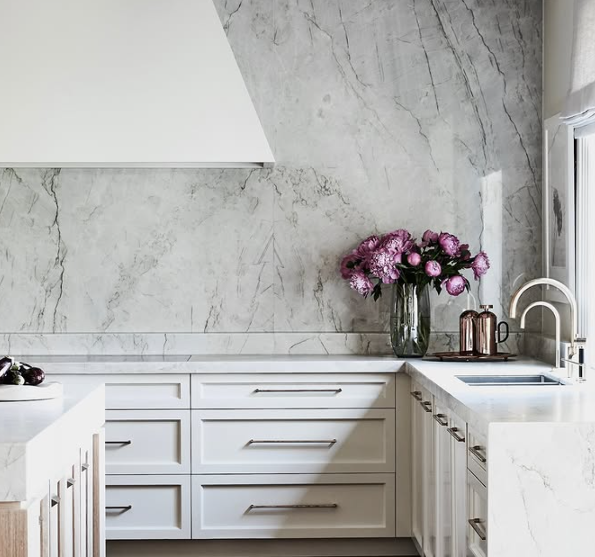 Modern kitchen with white cabinets, marble backsplash, and countertop. A vase with pink flowers and two copper mugs are on the counter near the window, which has a white frame.
