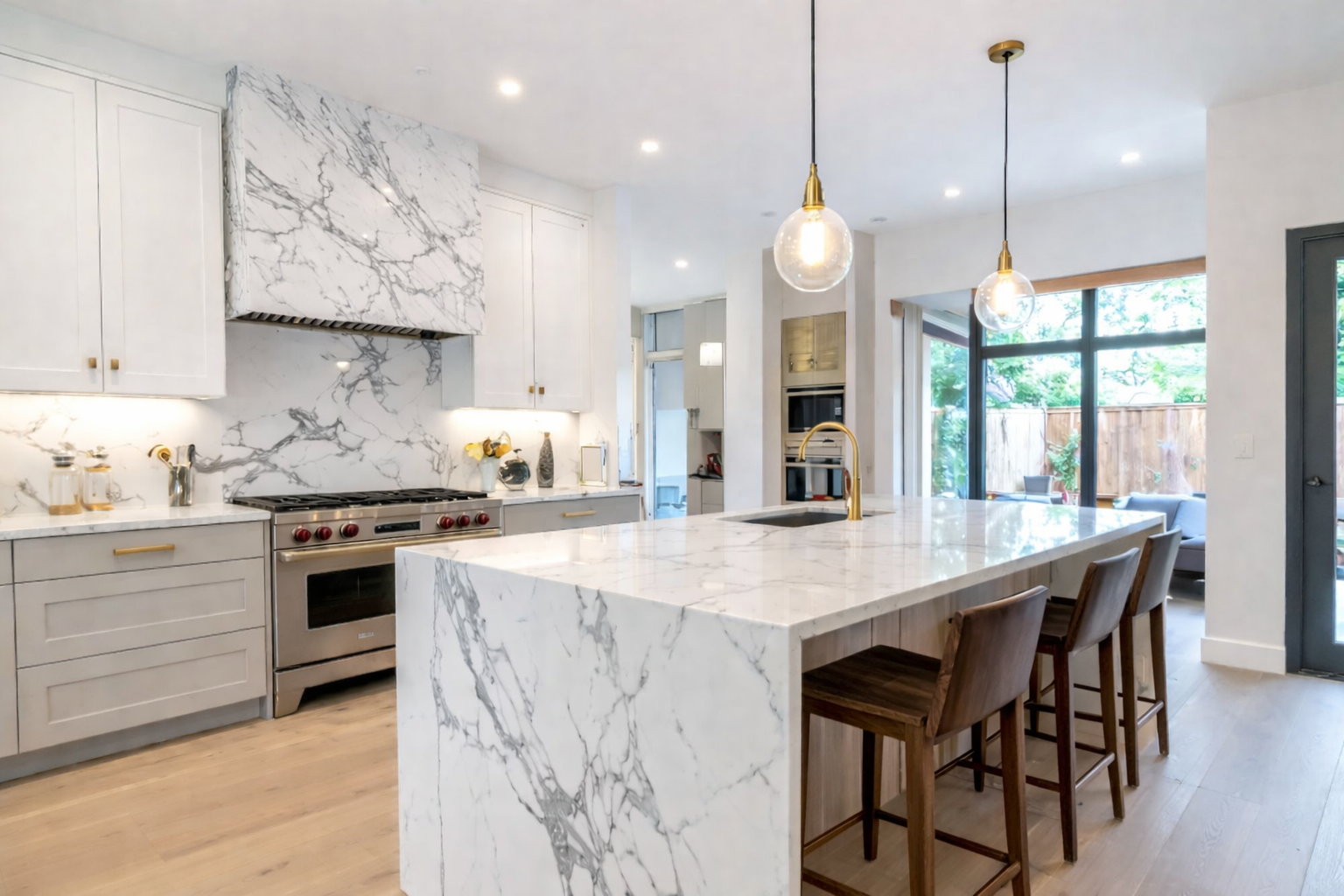 Modern kitchen with white marble countertops and backsplash, stainless steel stove, pendant lights, wooden chairs at island, and sliding glass doors leading to backyard with outdoor seating.