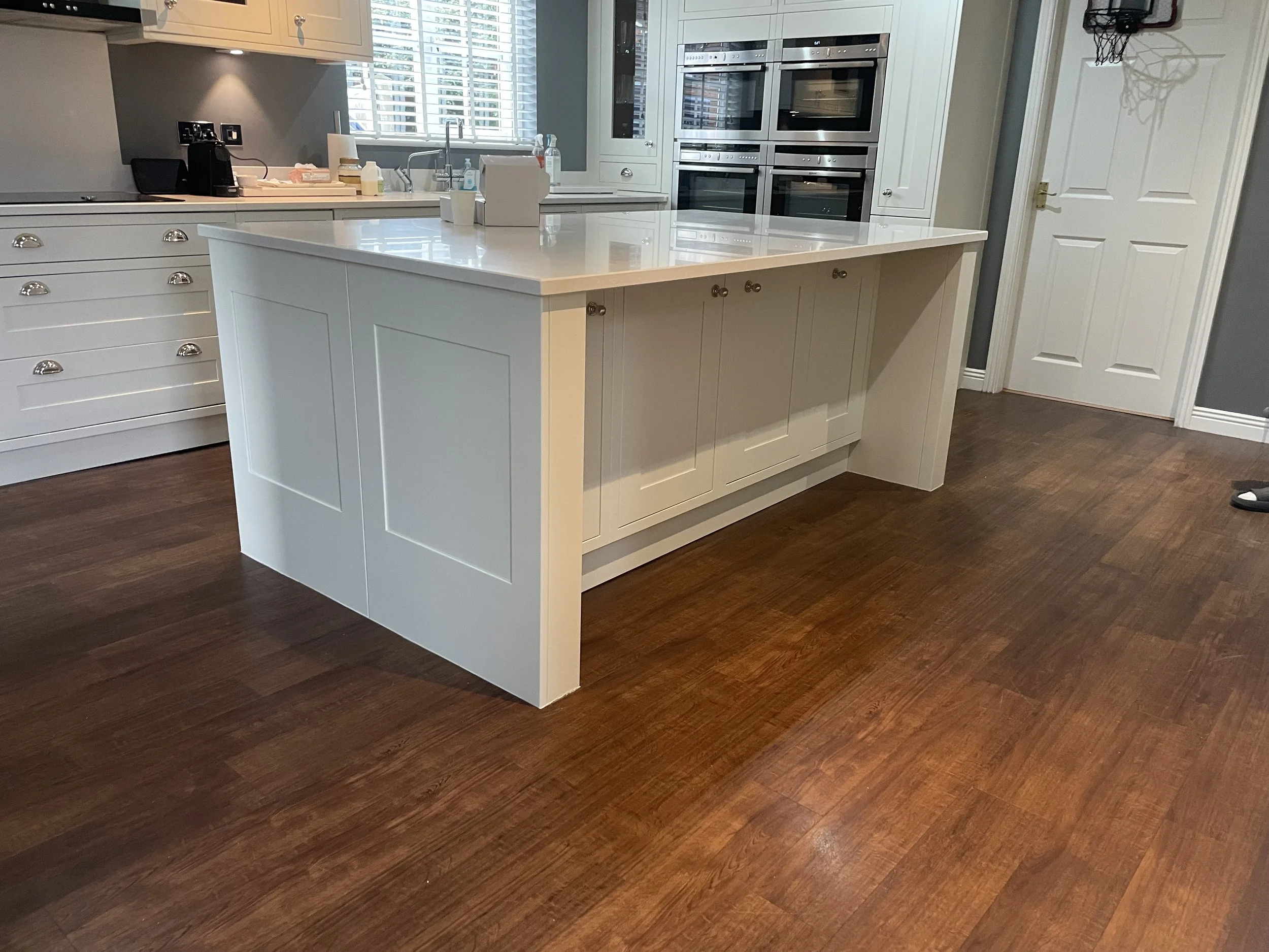 A kitchen with white cabinetry, a kitchen island, stainless steel double oven, coffee maker, and a window with white blinds.