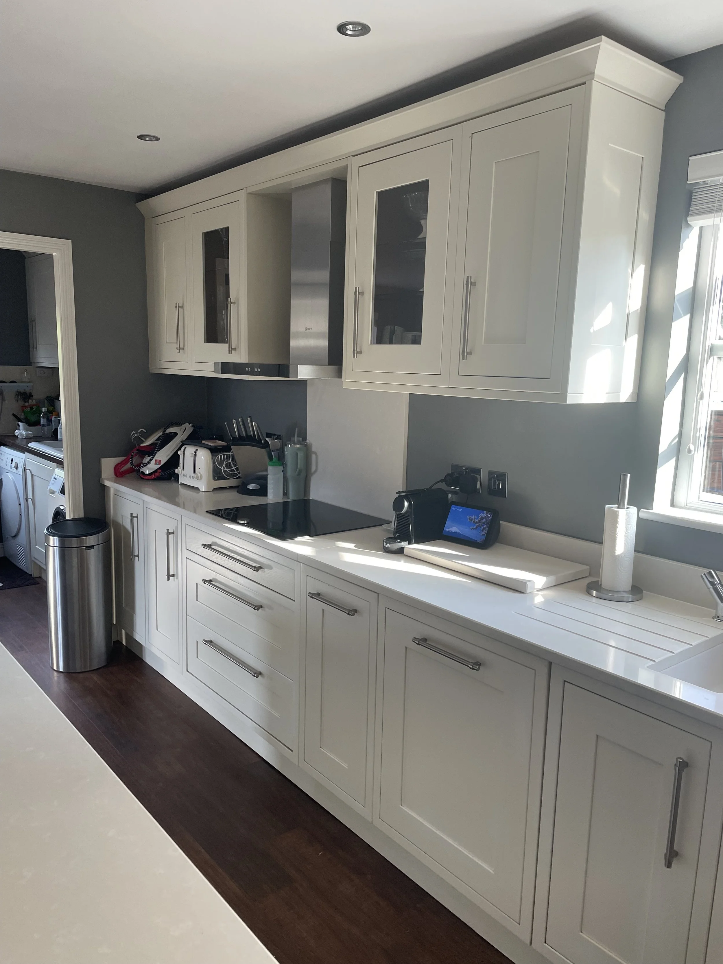 Modern kitchen with white cabinets, dark hardwood floor, stainless steel trash can, window with sunlight, and various small appliances on the countertop.