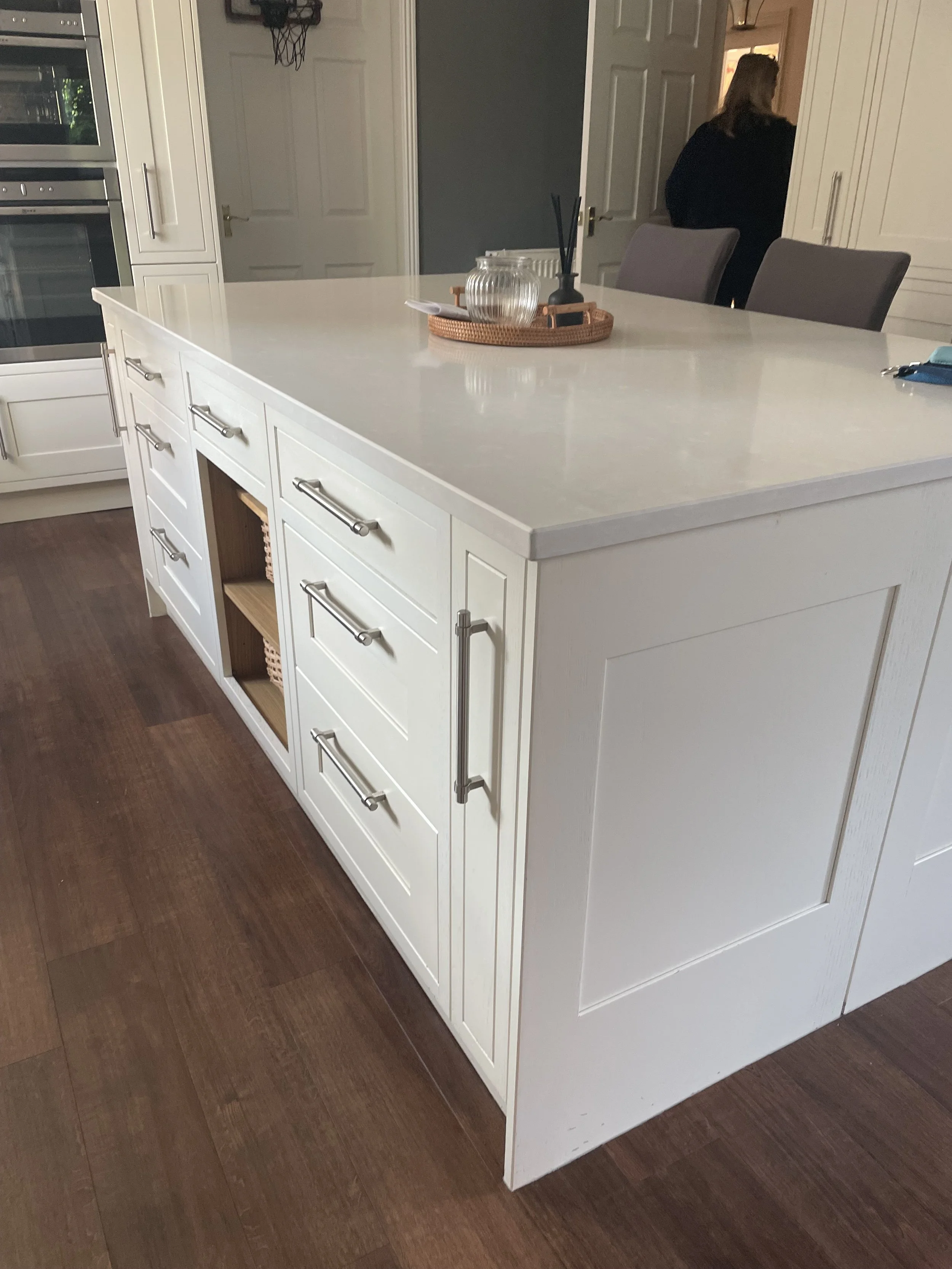 A white kitchen island with drawers and open shelves, a wooden countertop, with a tray on top holding glass jars and a black reed diffuser, and chairs in the background.