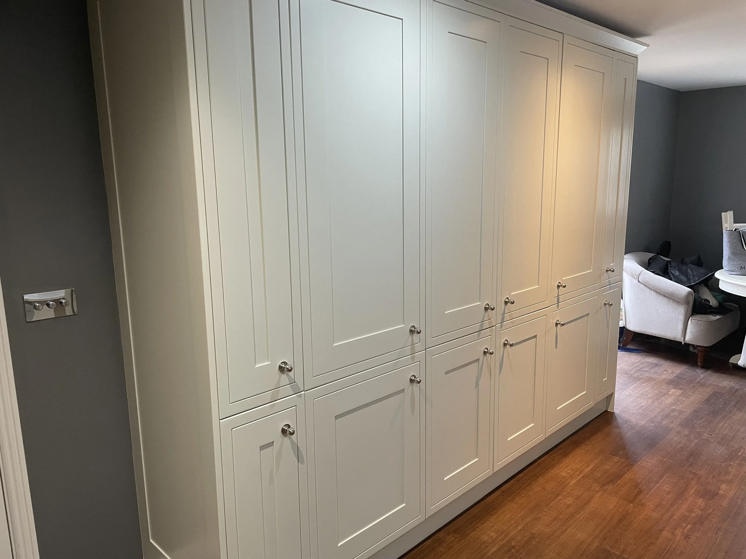 White built-in closet cabinets with silver knobs in a room with brown hardwood flooring and gray walls, partially visible furniture including an armchair and a side table.