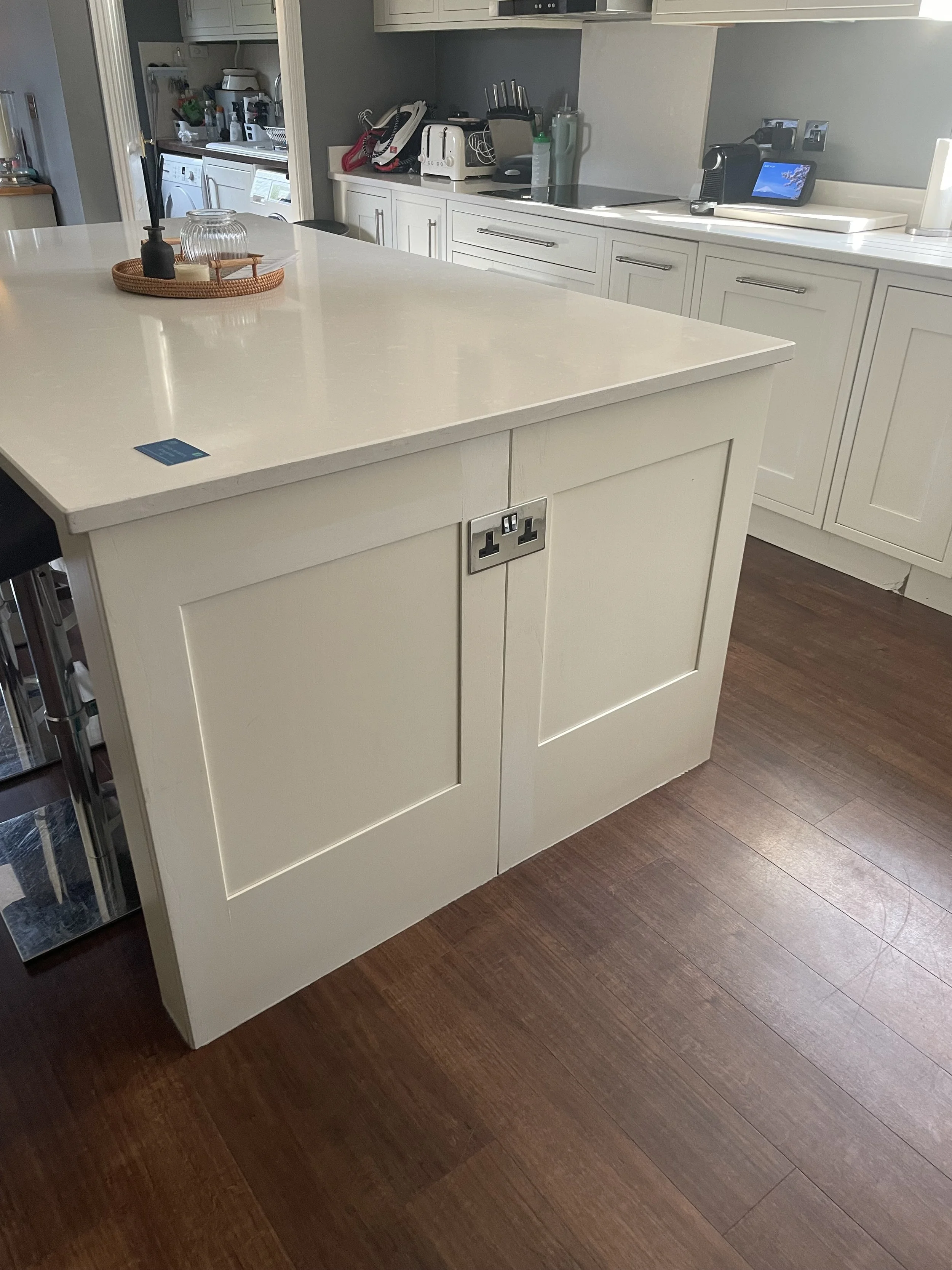 A kitchen island with a cream-colored base and a white countertop, with electrical outlets on the side. There are various kitchen appliances and utensils on the countertop in the background.