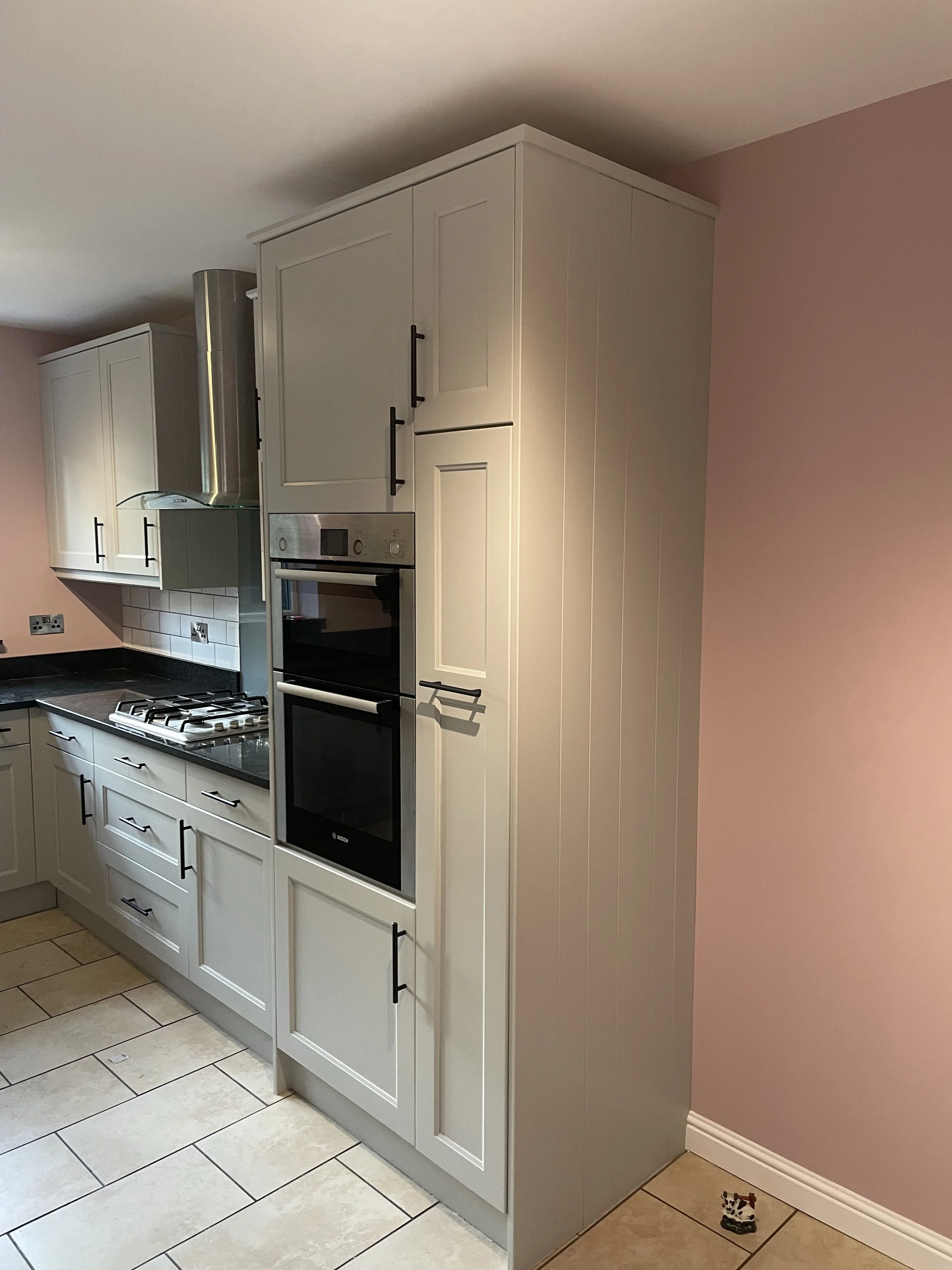 Kitchen with cream-colored cabinets, black handles, black countertops, a gas stove, an oven, a stainless steel range hood, pink walls, and tiled flooring.