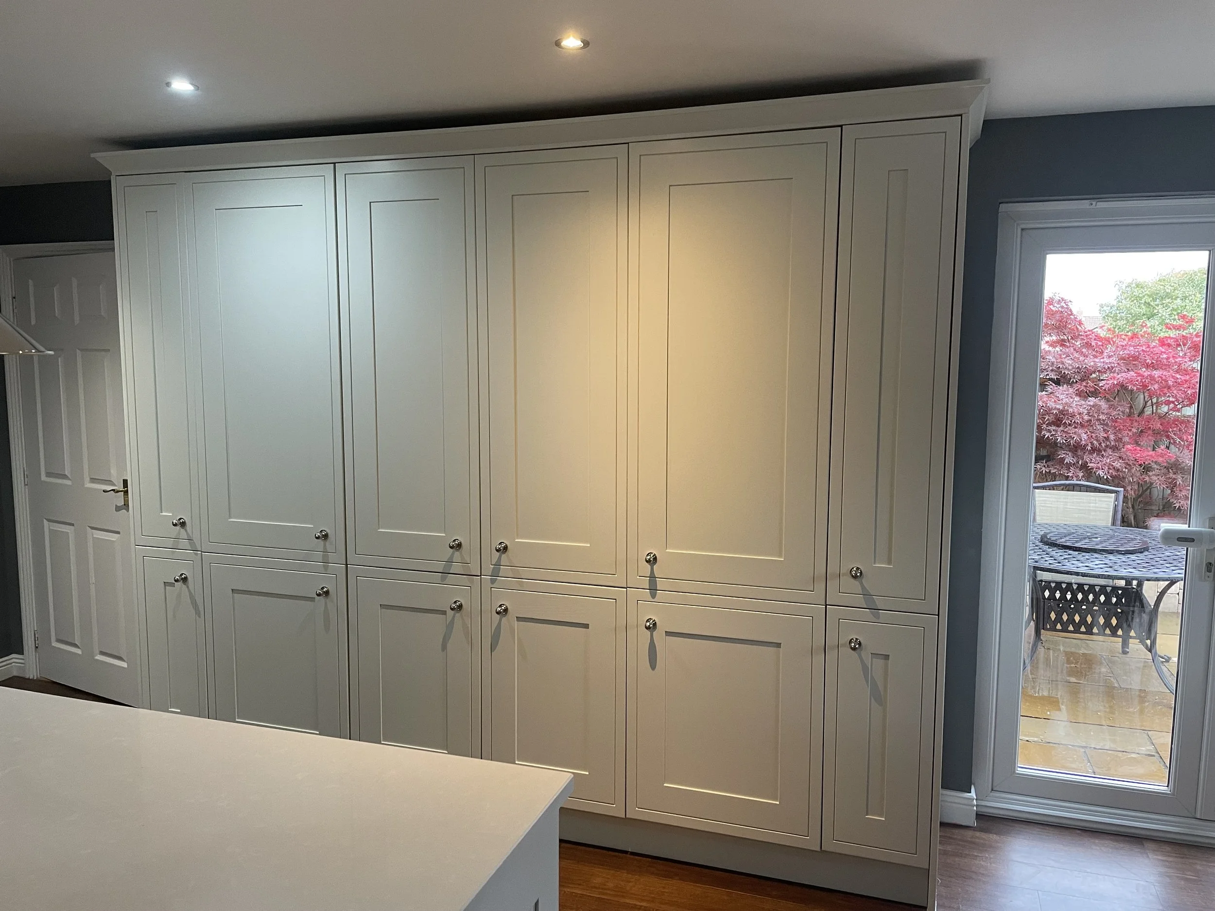 White kitchen cabinet with several doors and knobs next to a glass door leading to a patio with outdoor furniture and red trees.