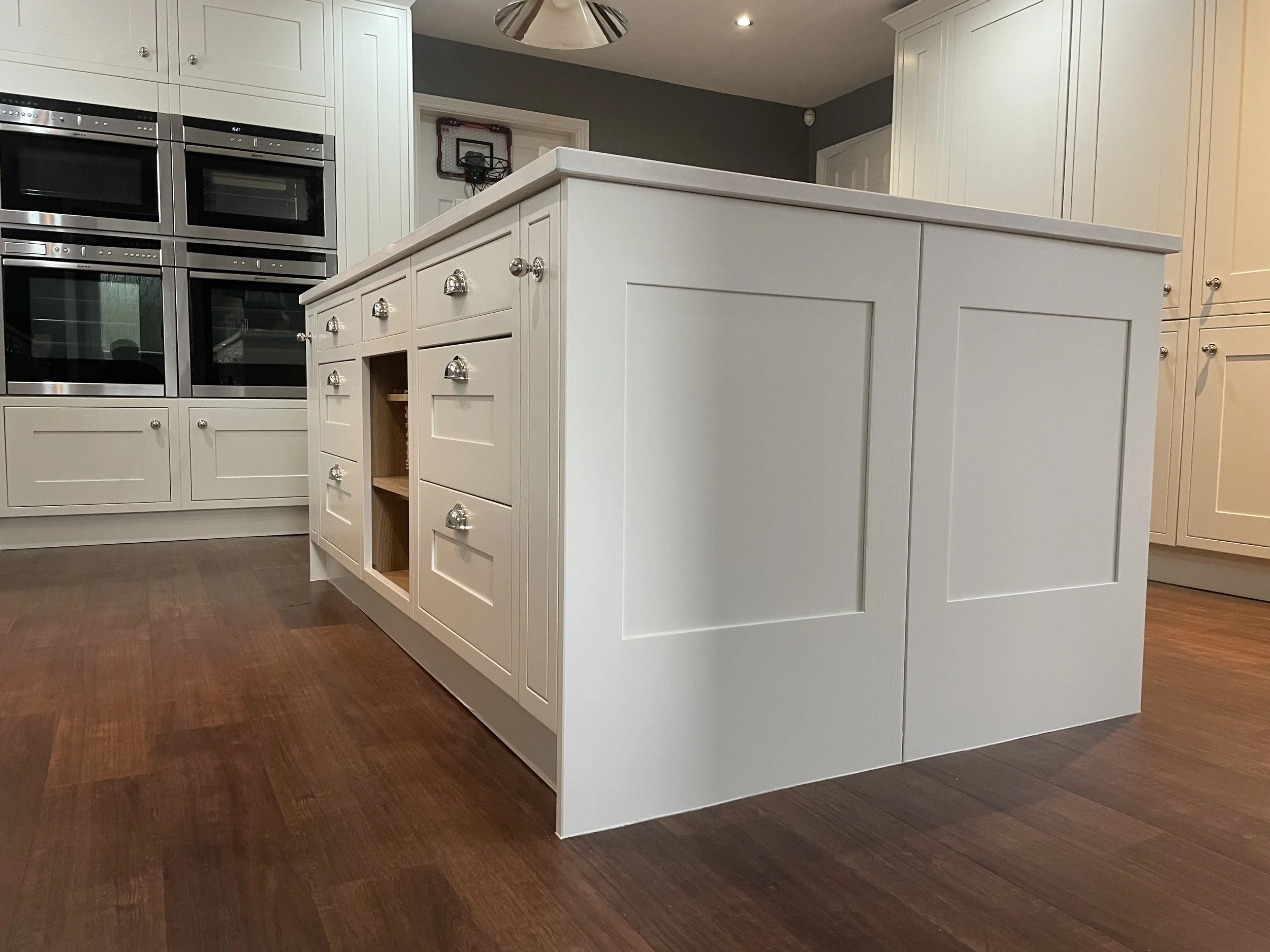 A modern white kitchen island with multiple drawers, cabinets, and two open shelves, situated on a hardwood floor. In the background, there are built-in cabinets and ovens.