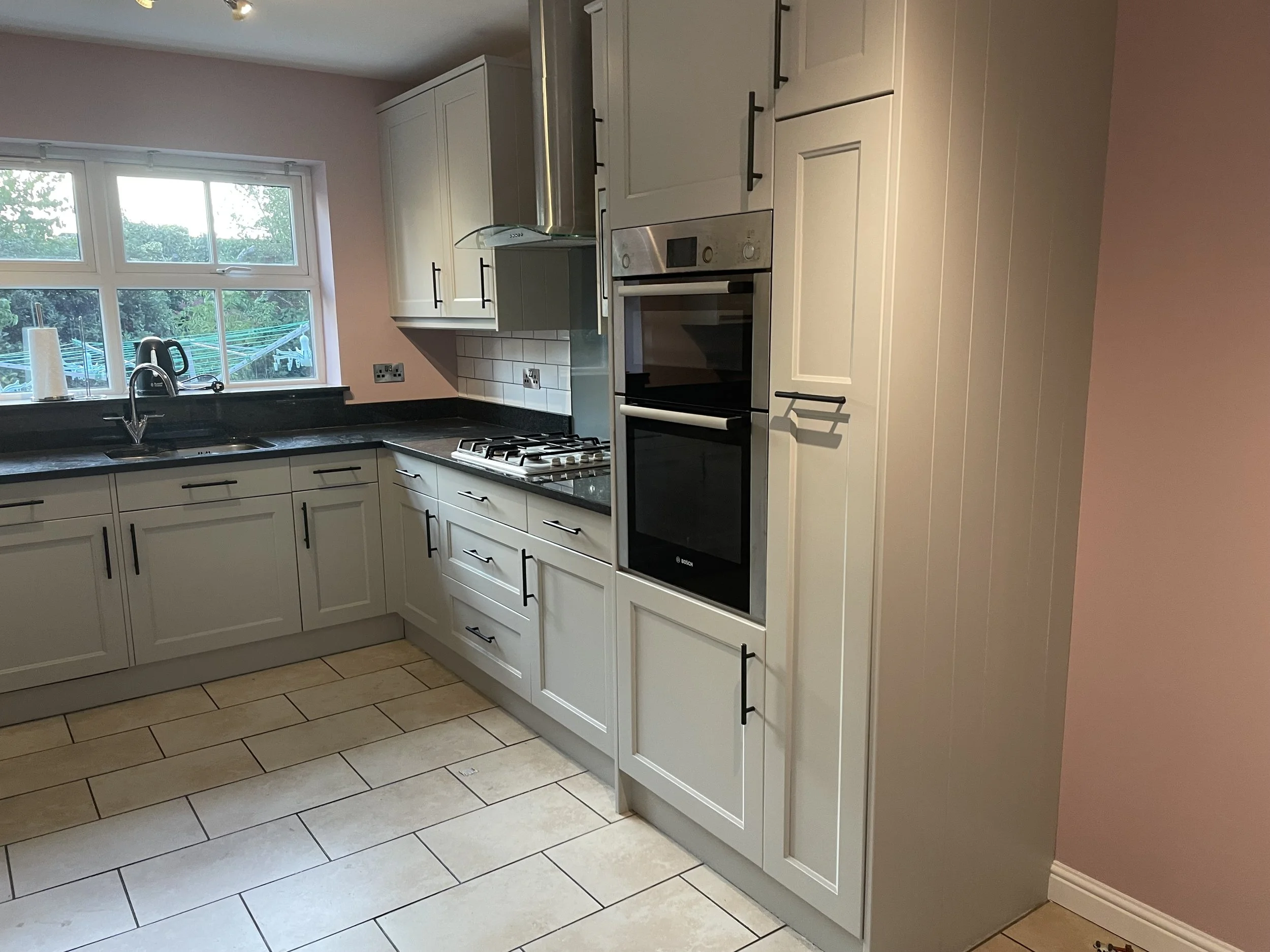 Interior view of a kitchen with white cabinets, black countertops, a window above the sink, a gas stove, an oven, and a pink wall on the right side.