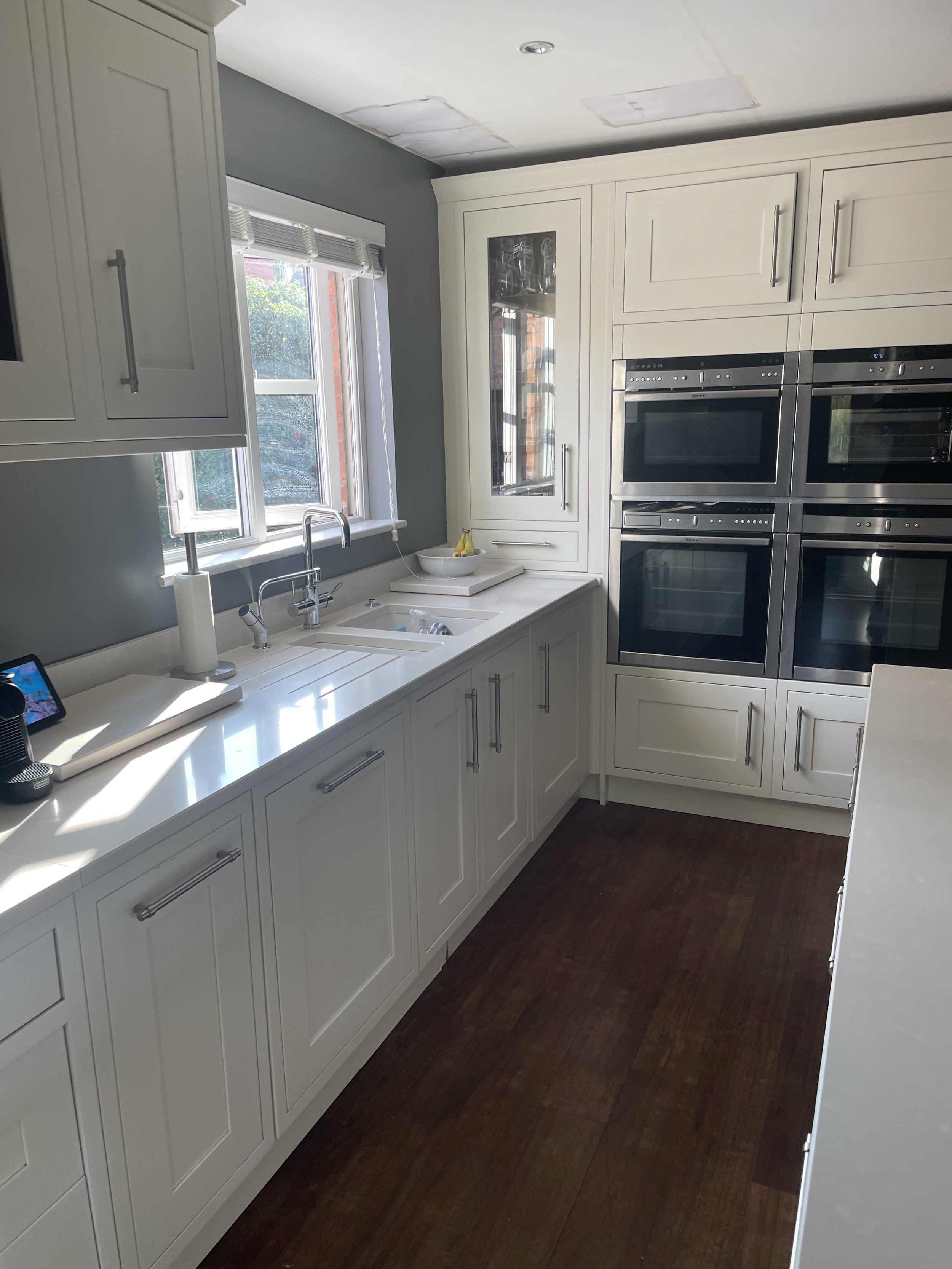 A modern kitchen with white cabinets, a window above the sink, and four stainless steel ovens stacked on the right wall. The dark wooden floor contrasts with the white cabinetry.