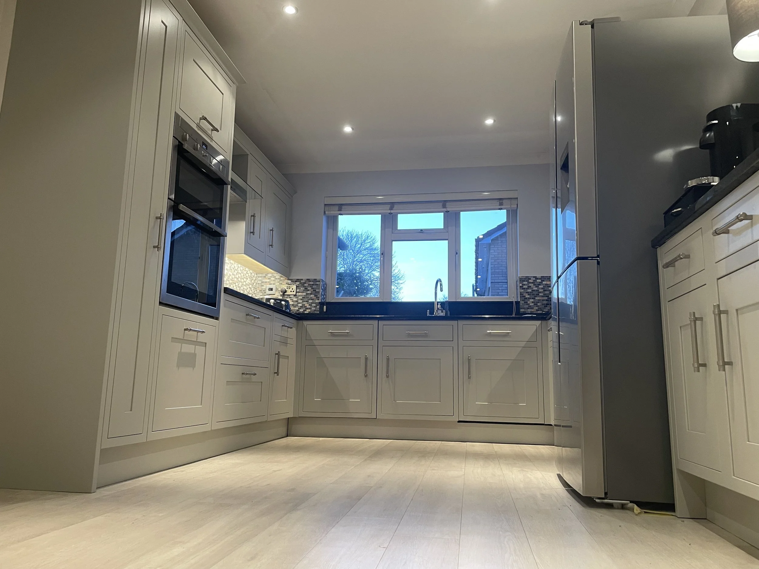 Modern kitchen with white cabinets, black countertops, stainless steel appliances, and a large window above the sink showing trees outside.