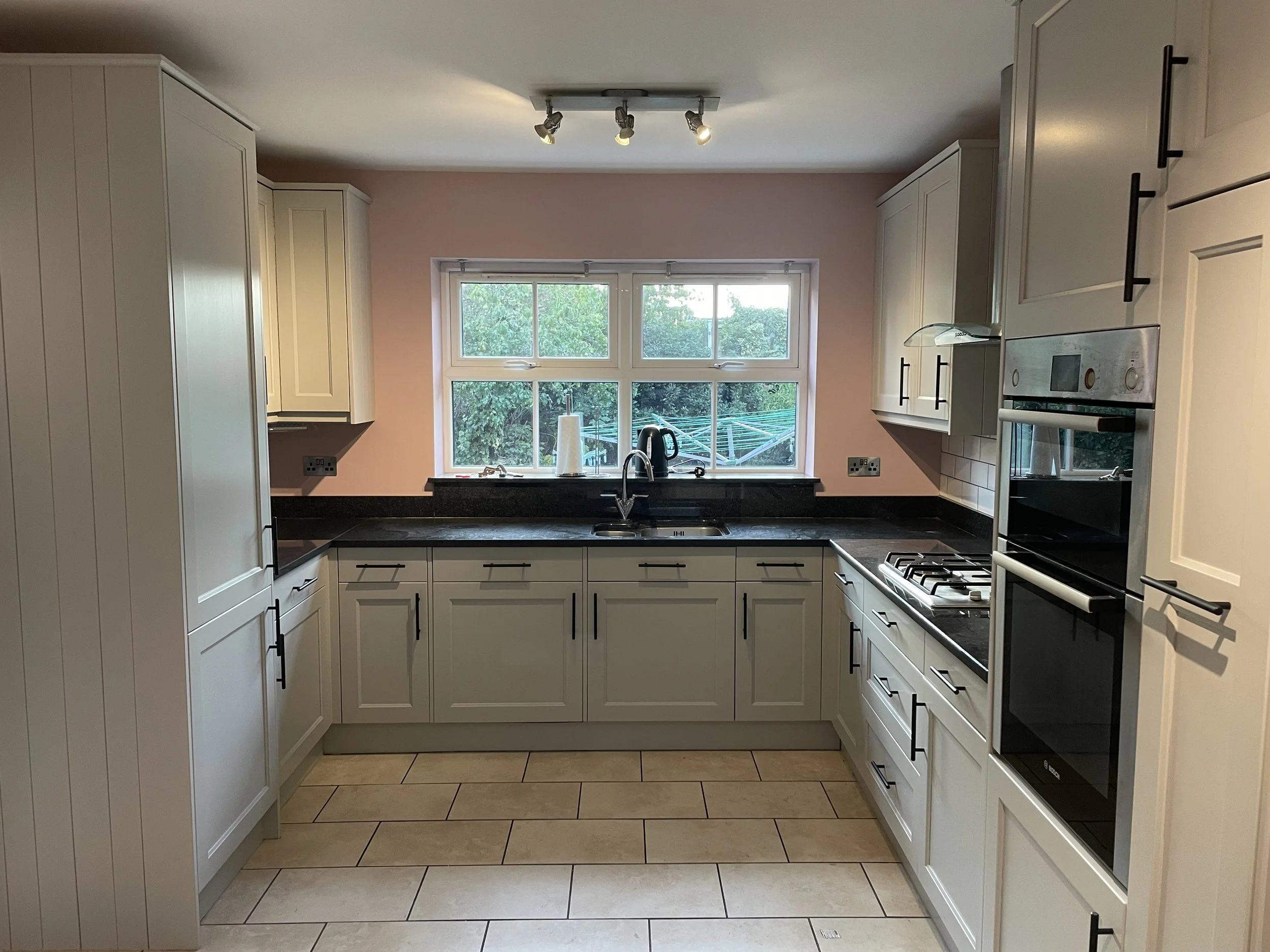 This is a kitchen with white cabinets, a black countertop, a sink beneath a window, and an oven and stove on the right side. There is a paper towel holder and an electric kettle near the window. The floor is tiled, and the walls are painted pink.