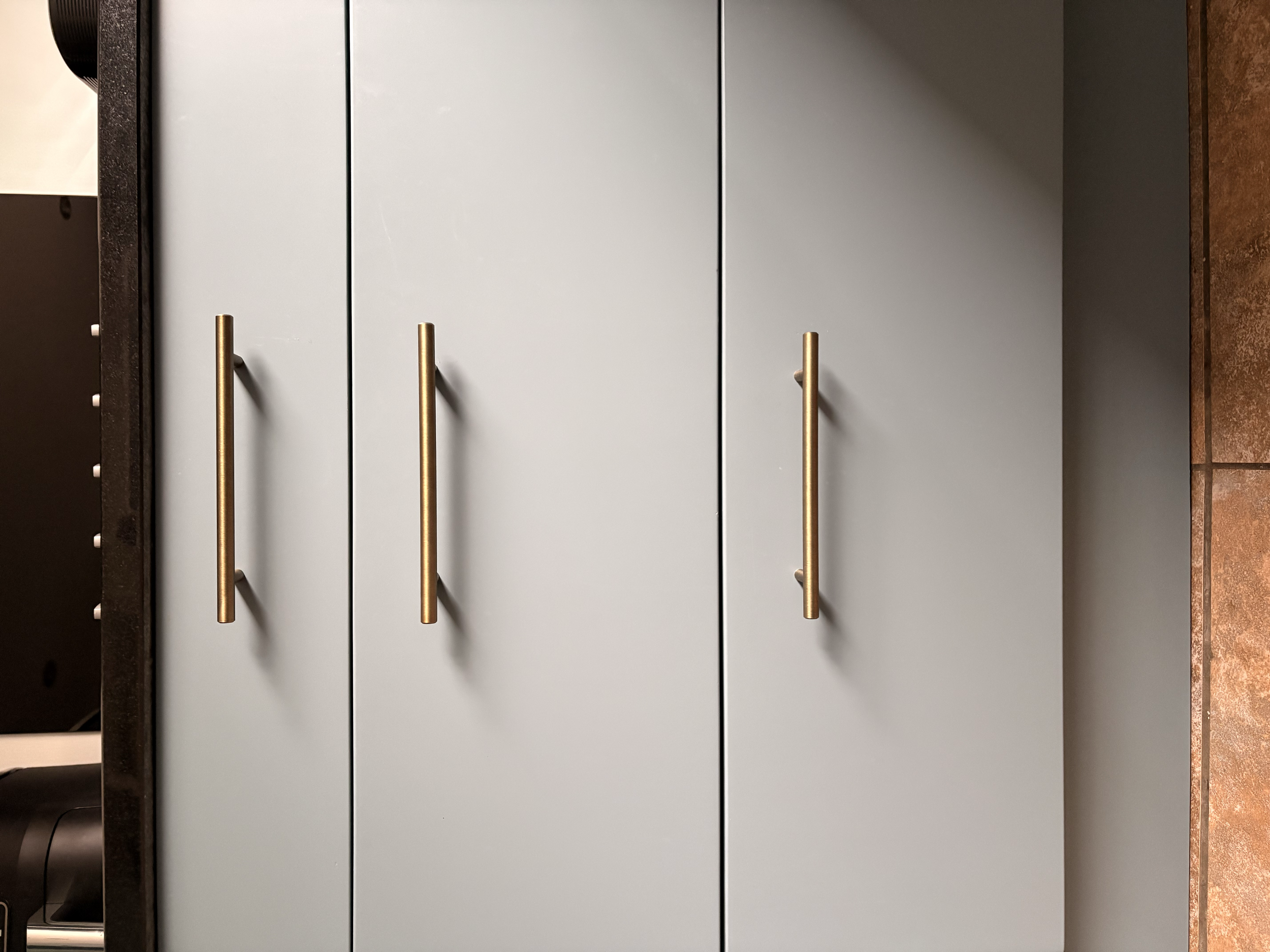 A light grey cabinet with three doors and long brass handles, situated next to a dark surface on the left and a brown tiled wall on the right.