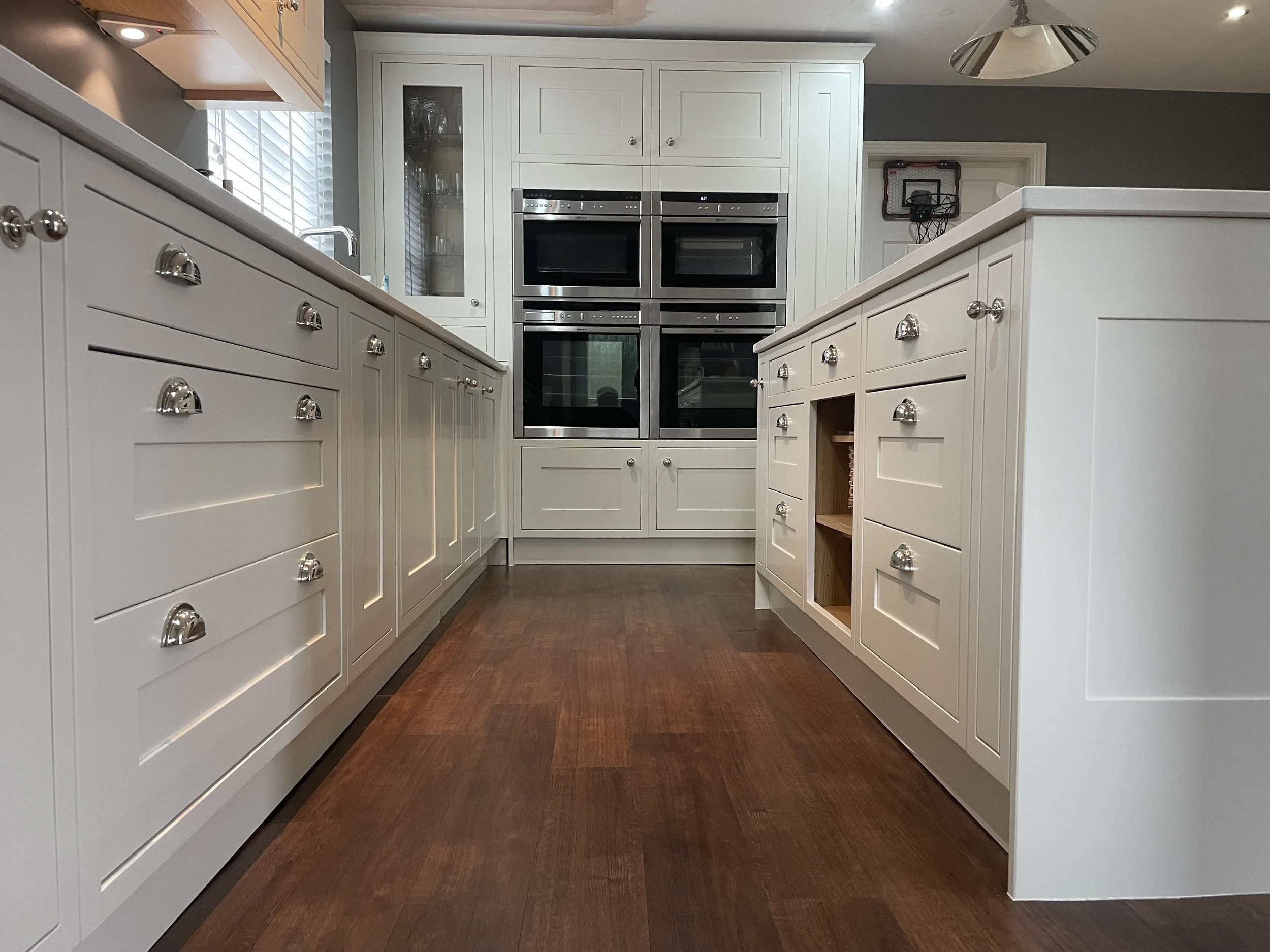 Modern white kitchen with multiple built-in stainless steel ovens and a center island with cabinetry, hardwood floor, and a window with blinds.