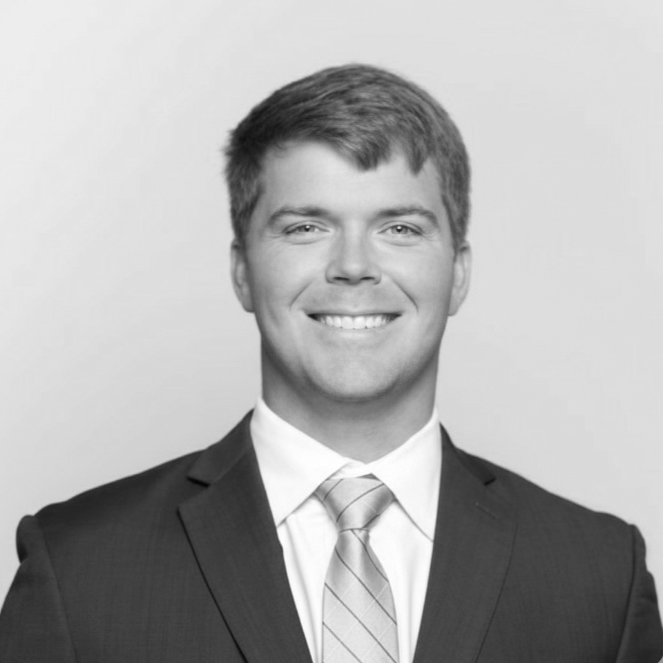 A smiling young man with light skin in a business suit, tie, and dress shirt posing against a plain background.