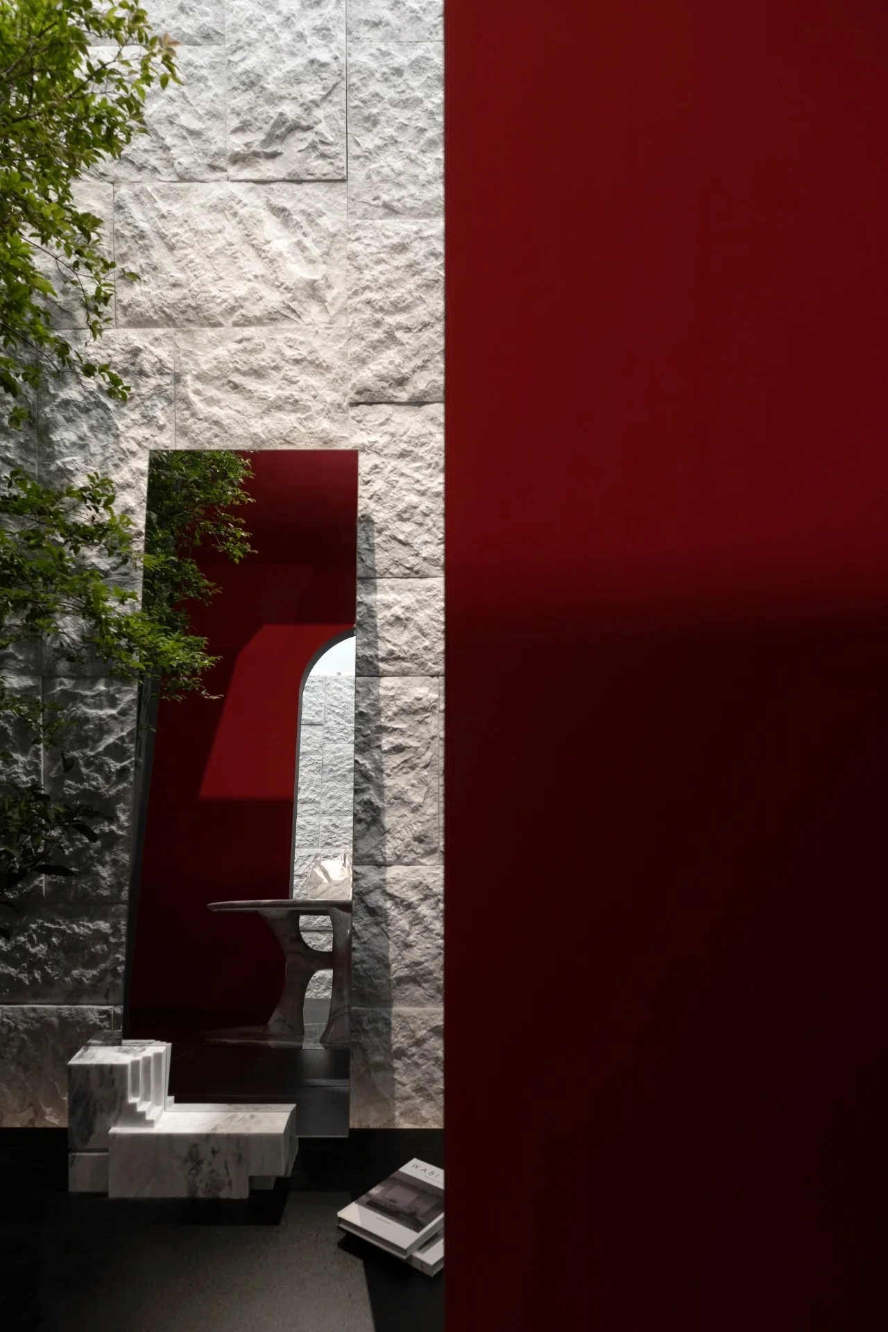Interior view of a modern space with a textured white stone wall, a narrow red wall, a small mirror reflecting a marble table, green trees outside the window, and books on the floor.