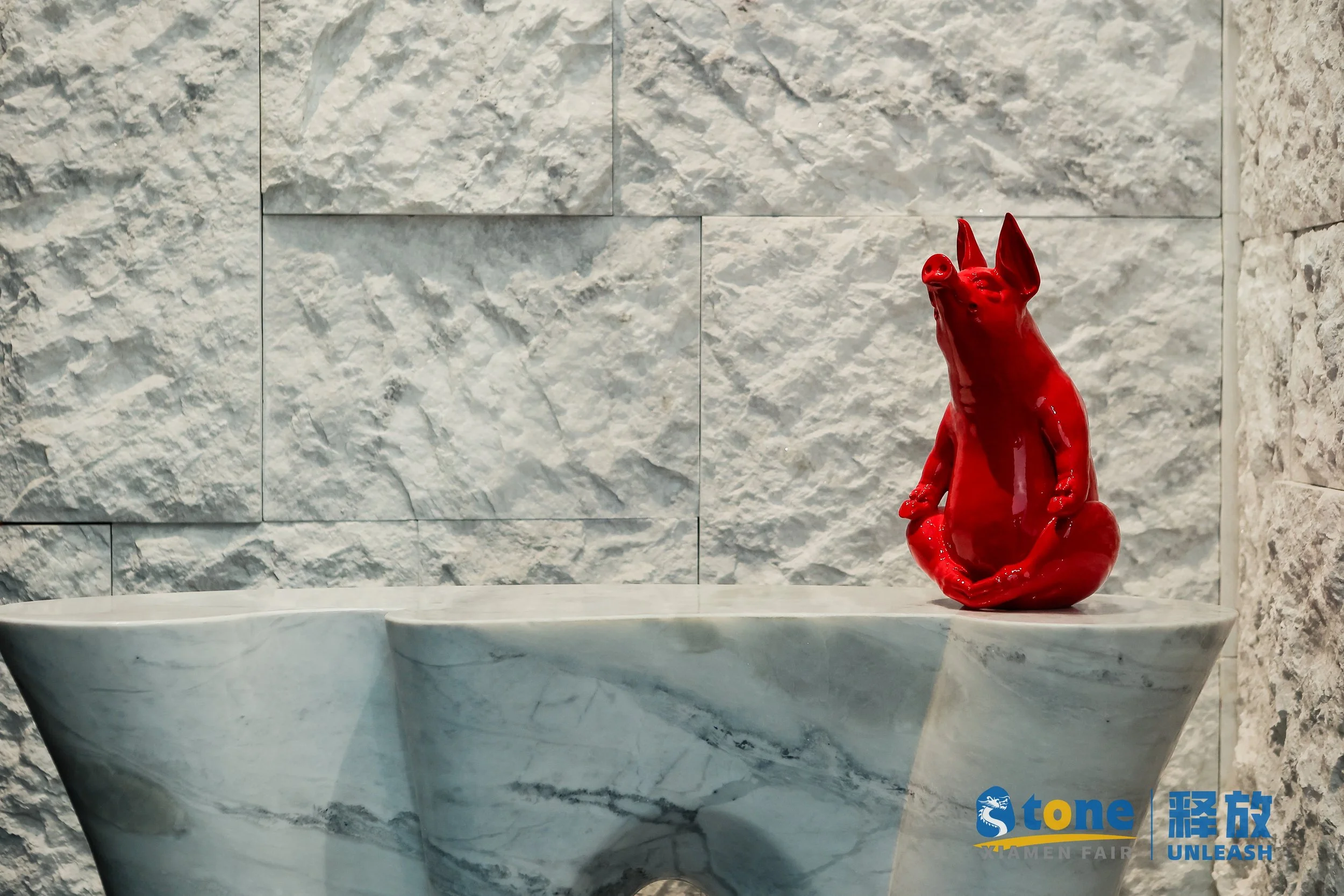A red decorative sculpture of a dog or wolf figure sitting on a marble surface against a textured stone wall background.