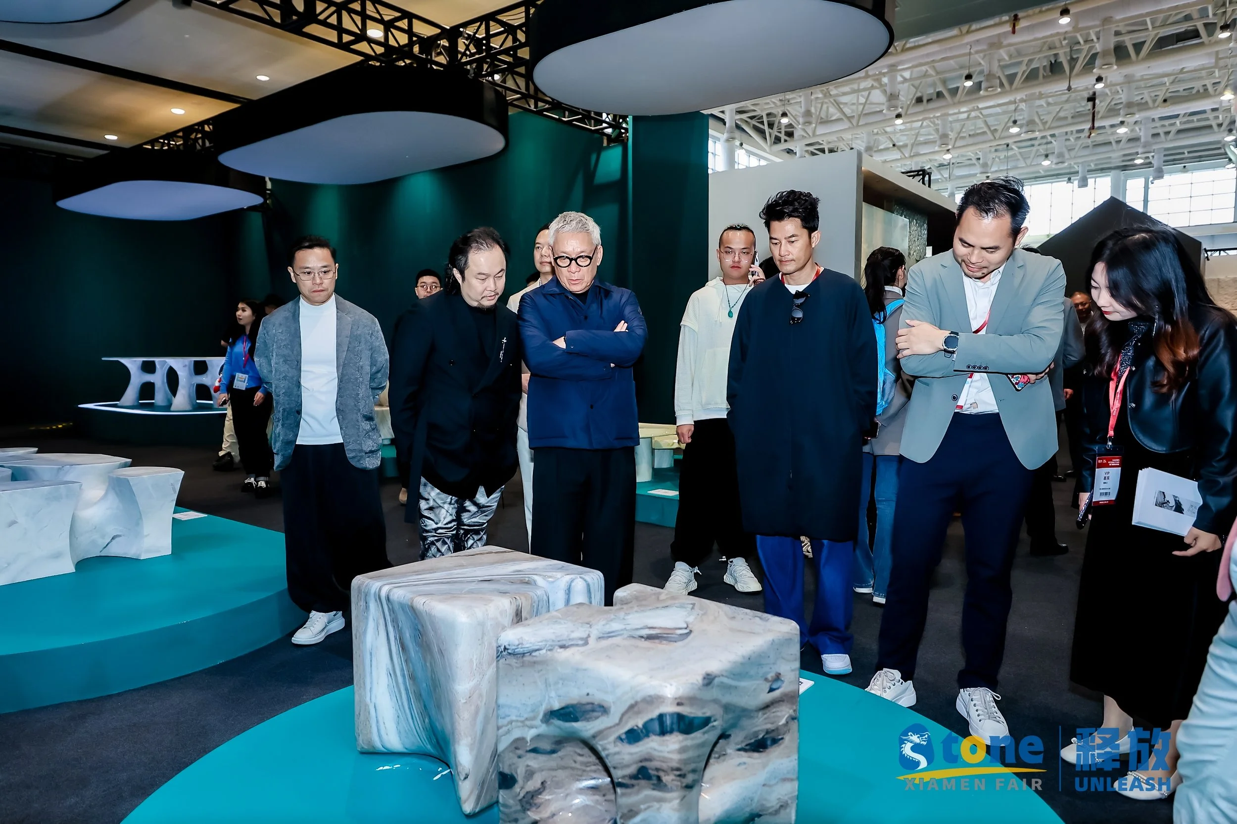 Group of people observing marble sculptures at an exhibition. The setting appears to be a modern gallery or trade show, with some individuals taking notes and examining the artworks.
