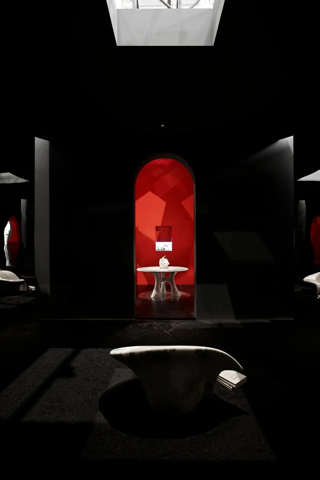 Interior of a dark museum room with a red-lit archway leading to a display with a white sculpture on a round table. Black walls with red accents and a white bench in the foreground.