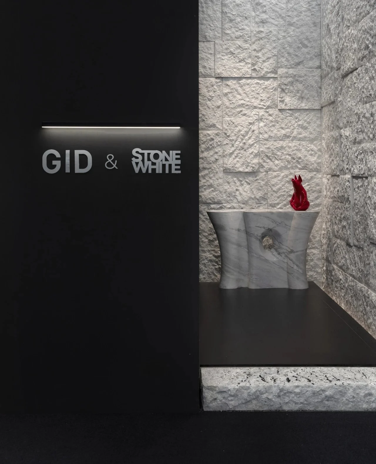 Interior view of a modern design store or gallery with black wall on the left displaying the names 'GID & Stone White' in white letters and a horizontal light strip above. On the right, there's a textured light stone wall with a white marble sculptur