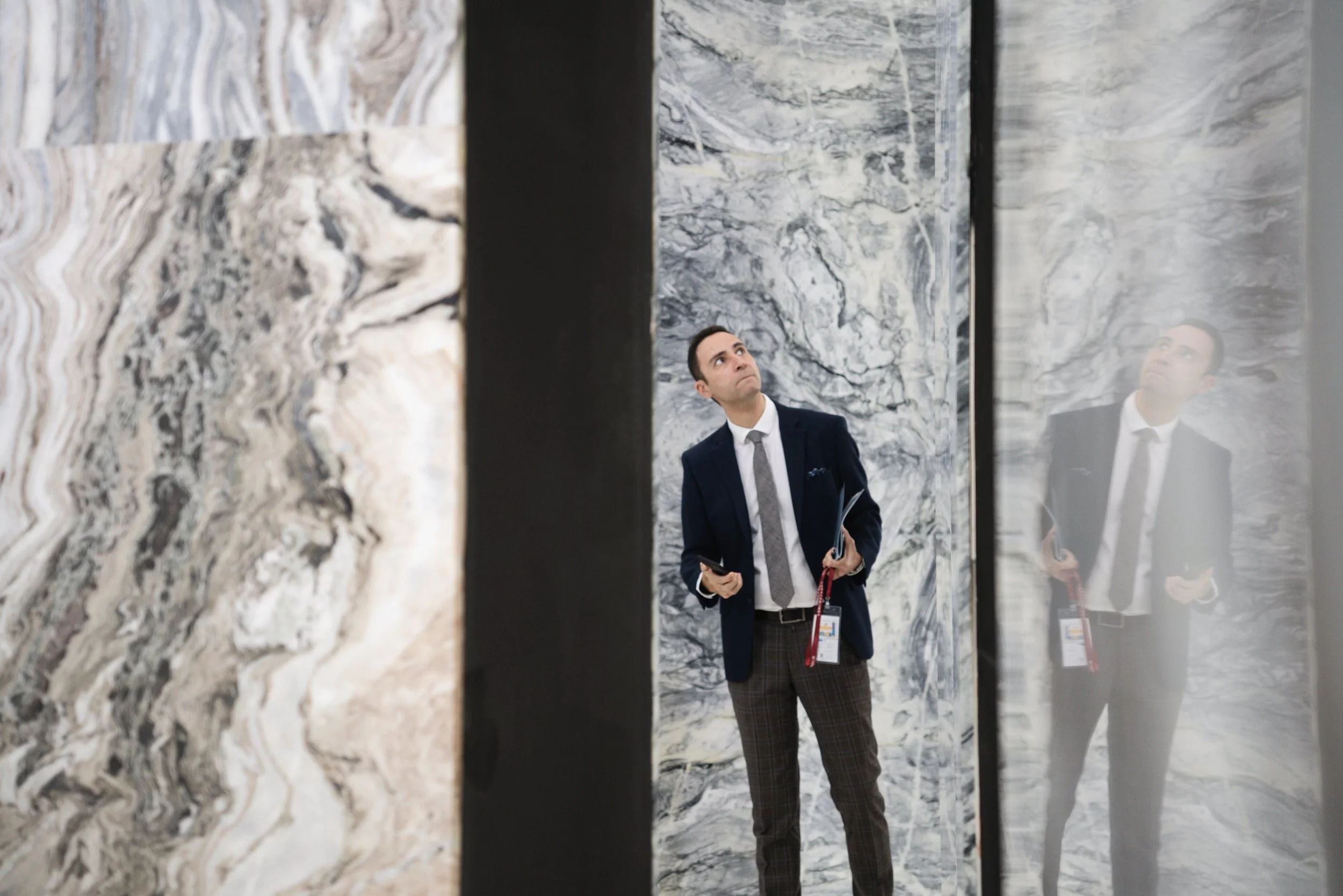 A man in business attire standing inside an elevator with marble walls, looking up and holding a phone and papers.