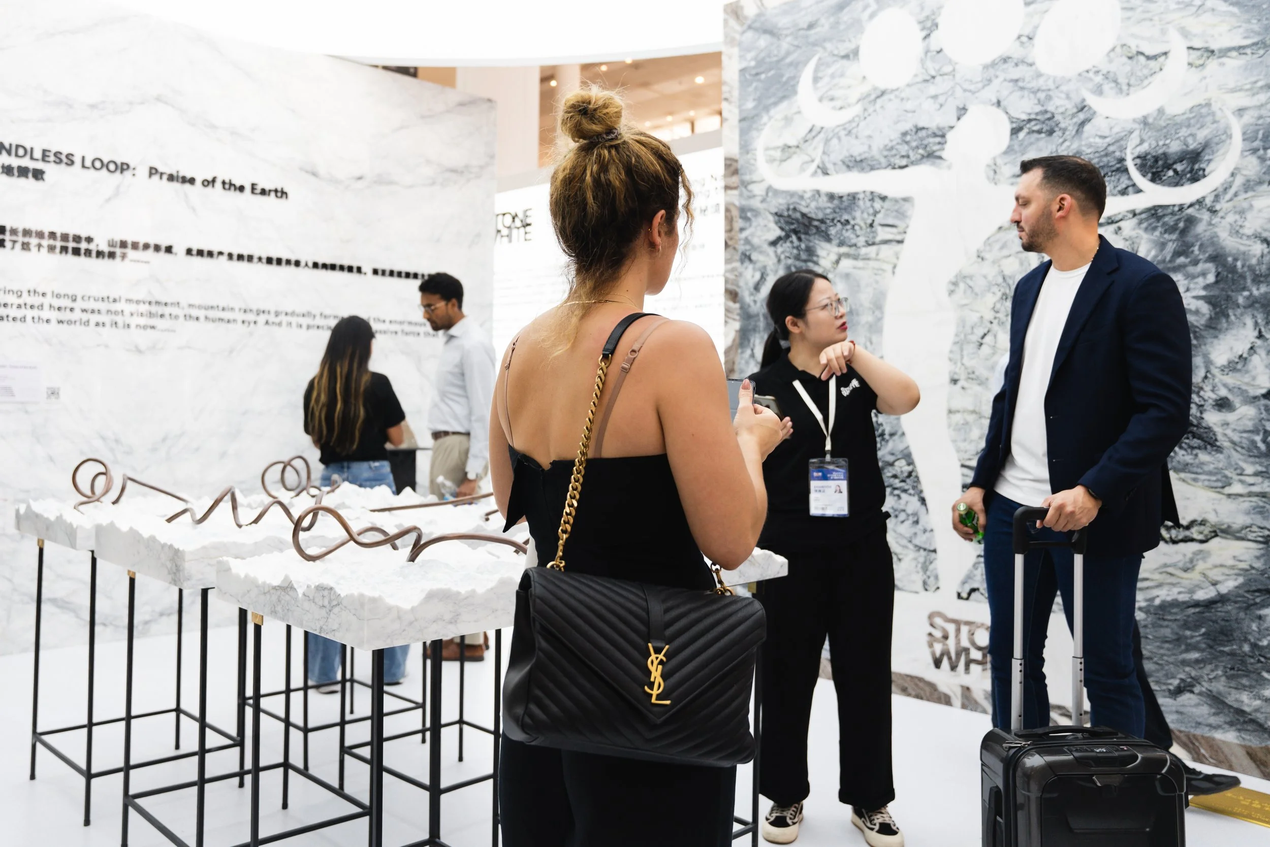 People at an exhibition talking to each other, with a display table and large marble-patterned wall in the background.