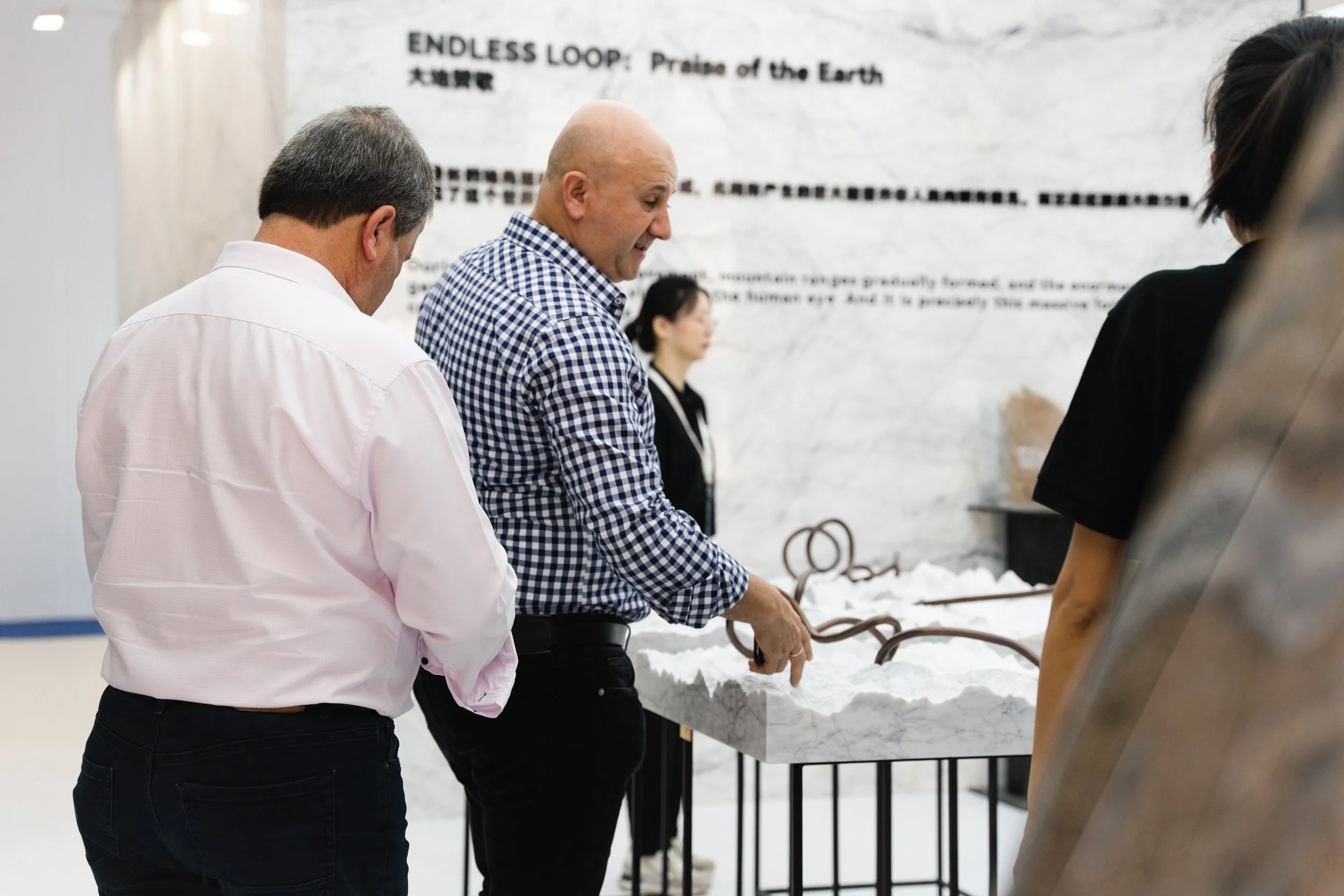 Two men are observing a marble sculpture titled 'Endless Loop: Praise of the Earth' at an art exhibit, with a woman in black in the background.