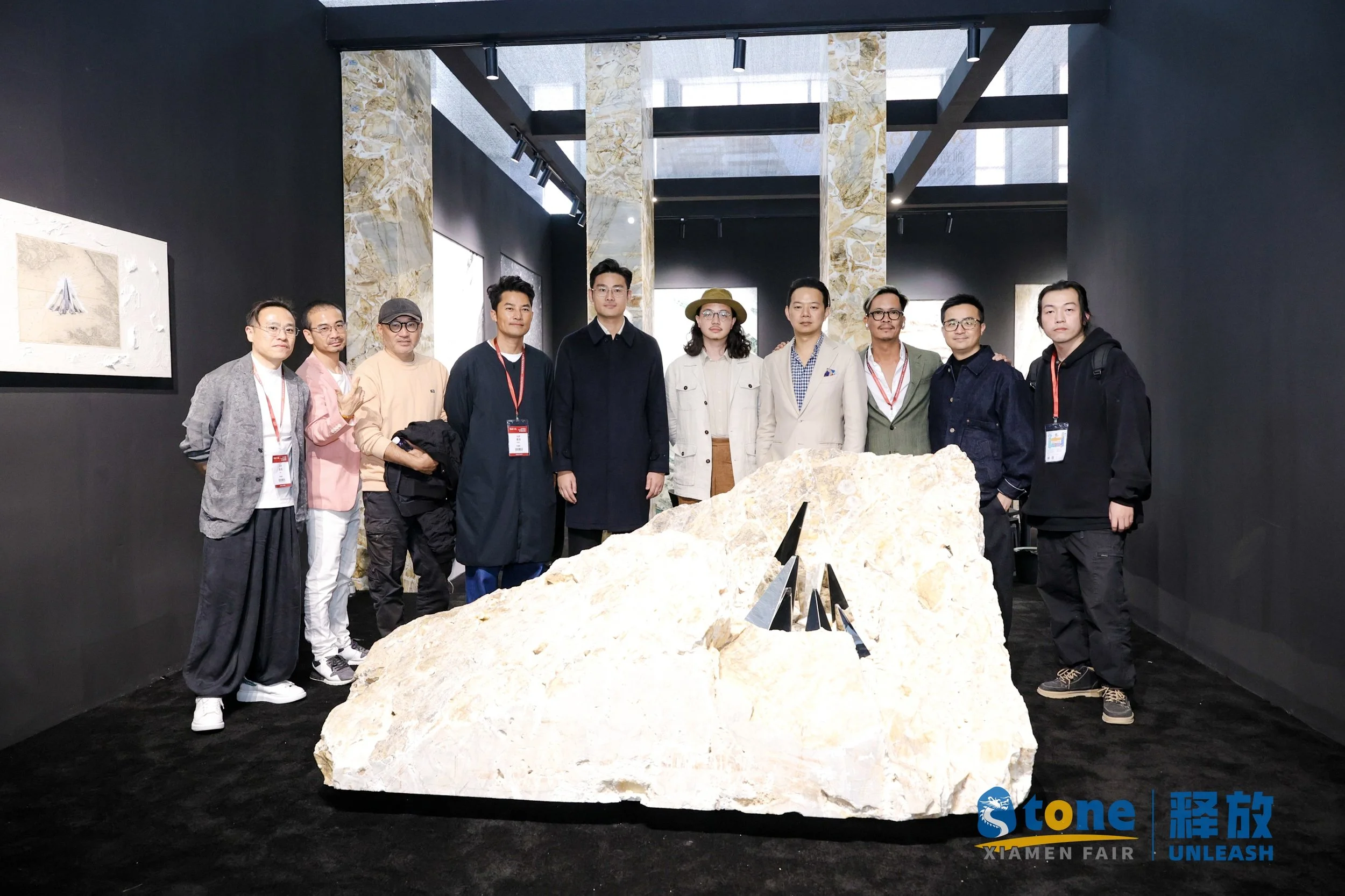 A group of ten people standing behind a large beige rock with black triangular sculptures on it at an art exhibition named Xiamen Fair. The background has a dark wall with artworks and a marble pillar. The group is posing for a photo.