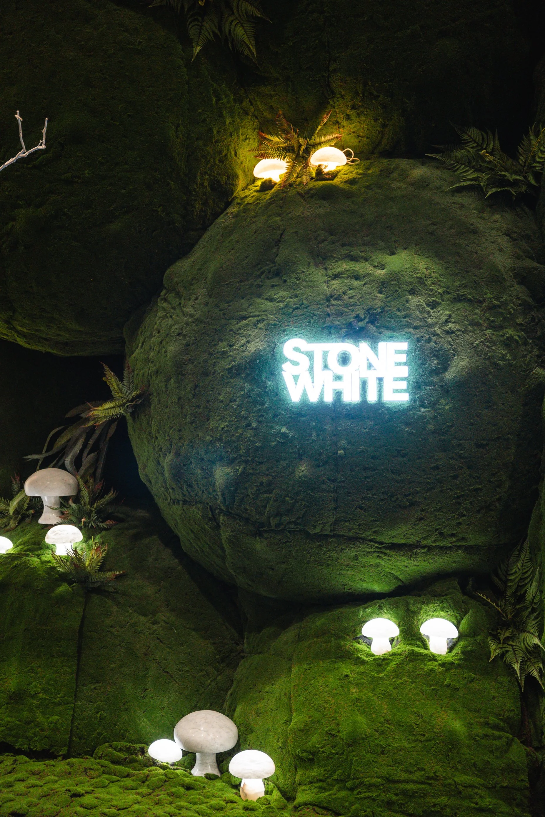 Green moss-covered rocks with illuminated white mushroom sculptures and a neon sign that says "Stone White."