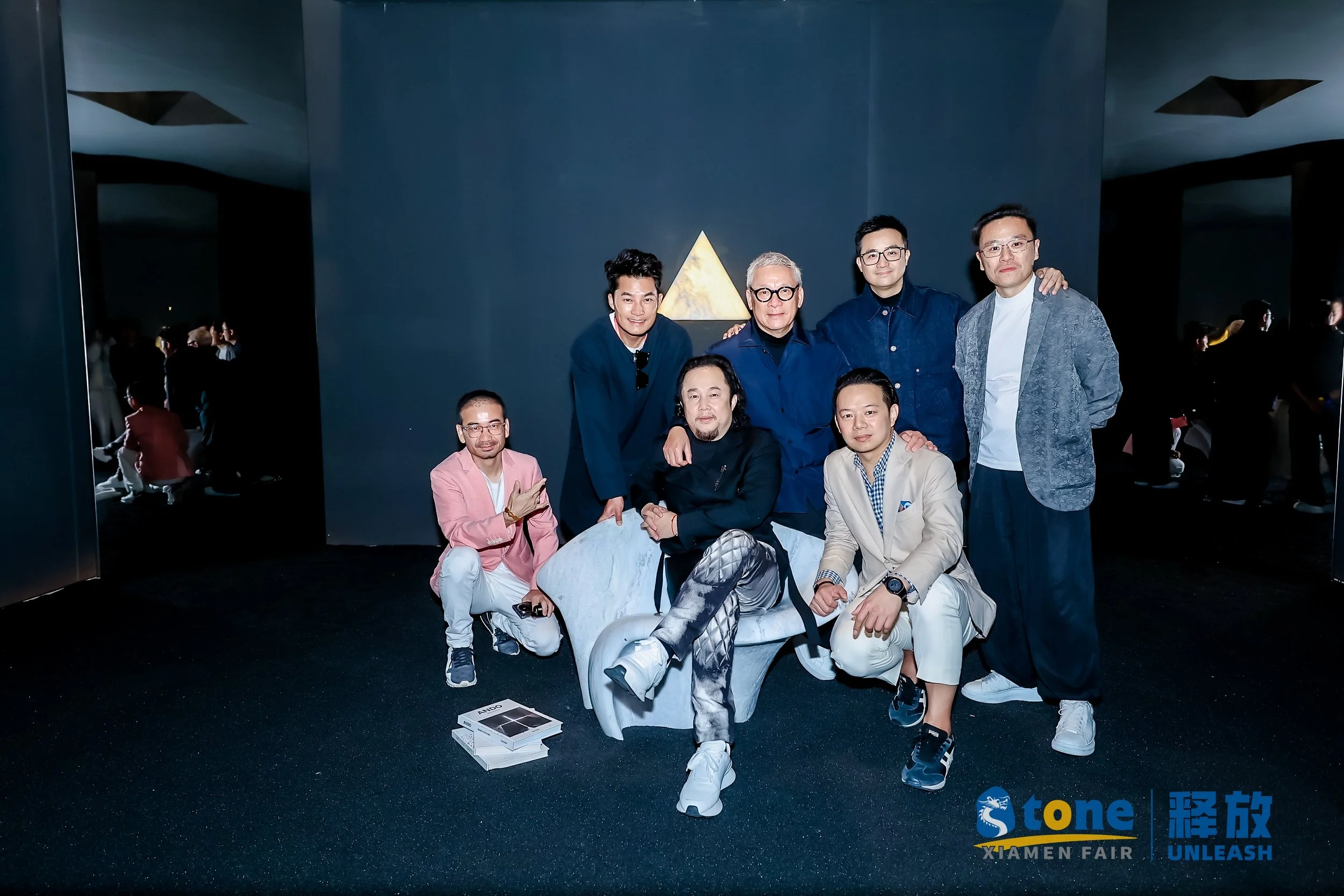 A group of nine men posing together at the Xiamen Fair, with some kneeling and others standing, in front of a dark blue wall with a golden triangle symbol. The bottom right corner has the logos for 'Stone,' 'Xiamen Fair,' and 'Unleash.'