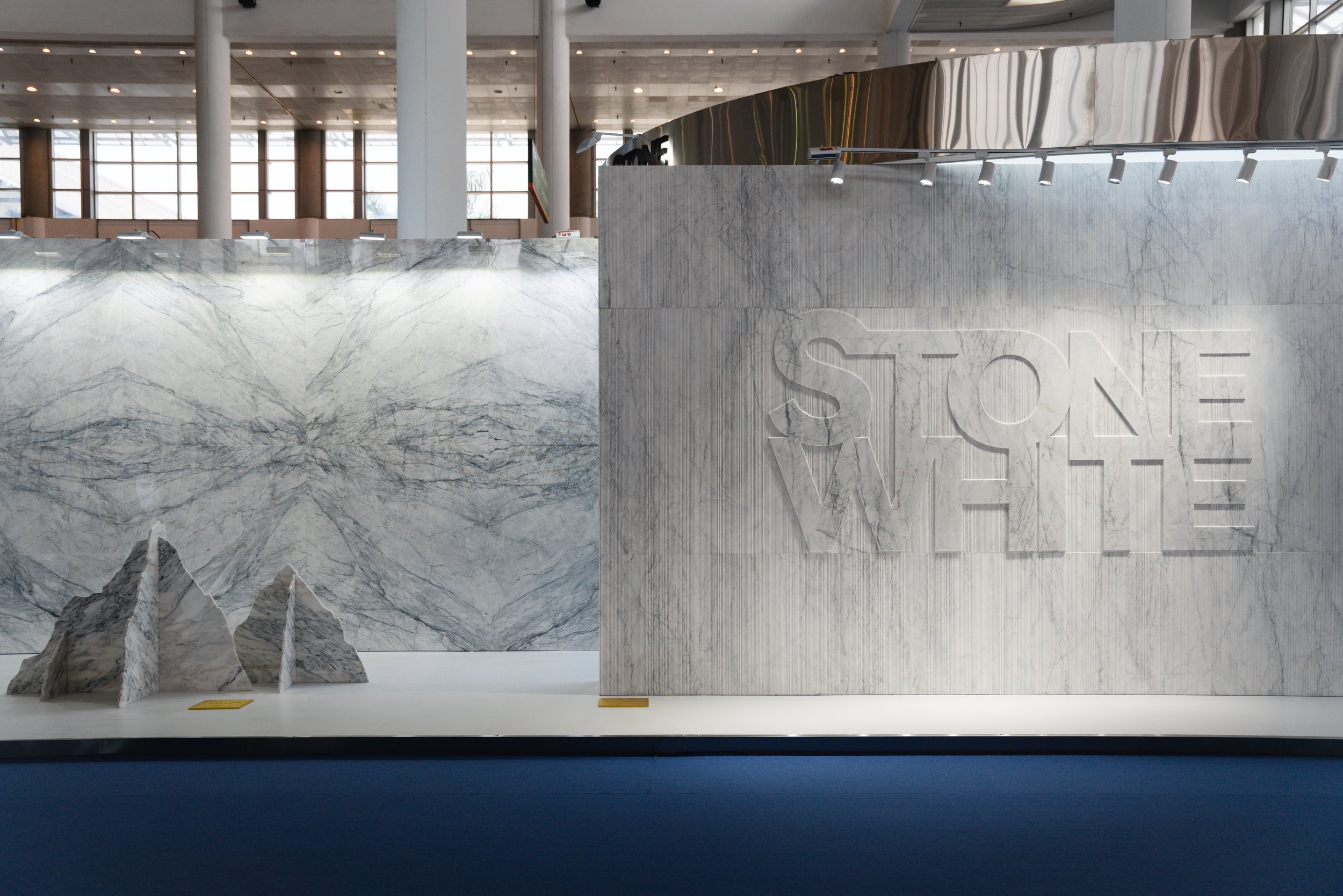 Interior of a building showcasing large marble display with the words 'Stone White' engraved on it, marble rocks displayed on the left, and architectural displays on the upper part of the wall.