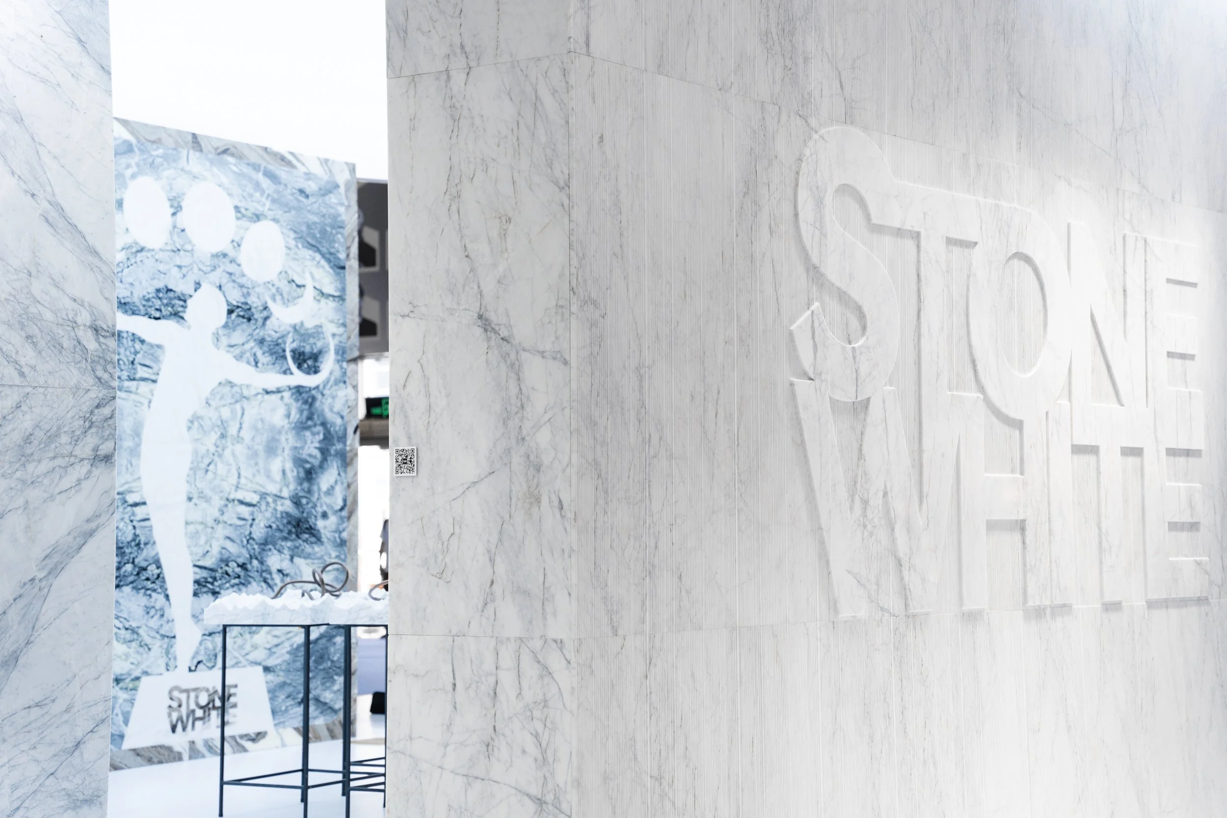 Marble wall with embossed 'STONE WHITE' logo and a distant poster featuring a figure holding five balloons, with 'STONE WHITE' text and a QR code.