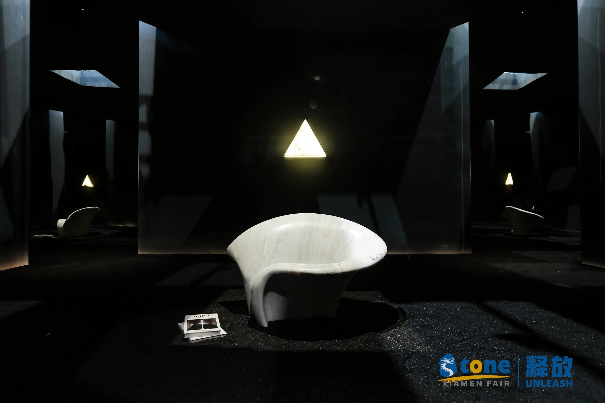 Modern exhibition space with a sculptural white chair in the foreground, dark walls, and triangular illuminated art pieces in the background, with a brochure and corporate logos on the floor.
