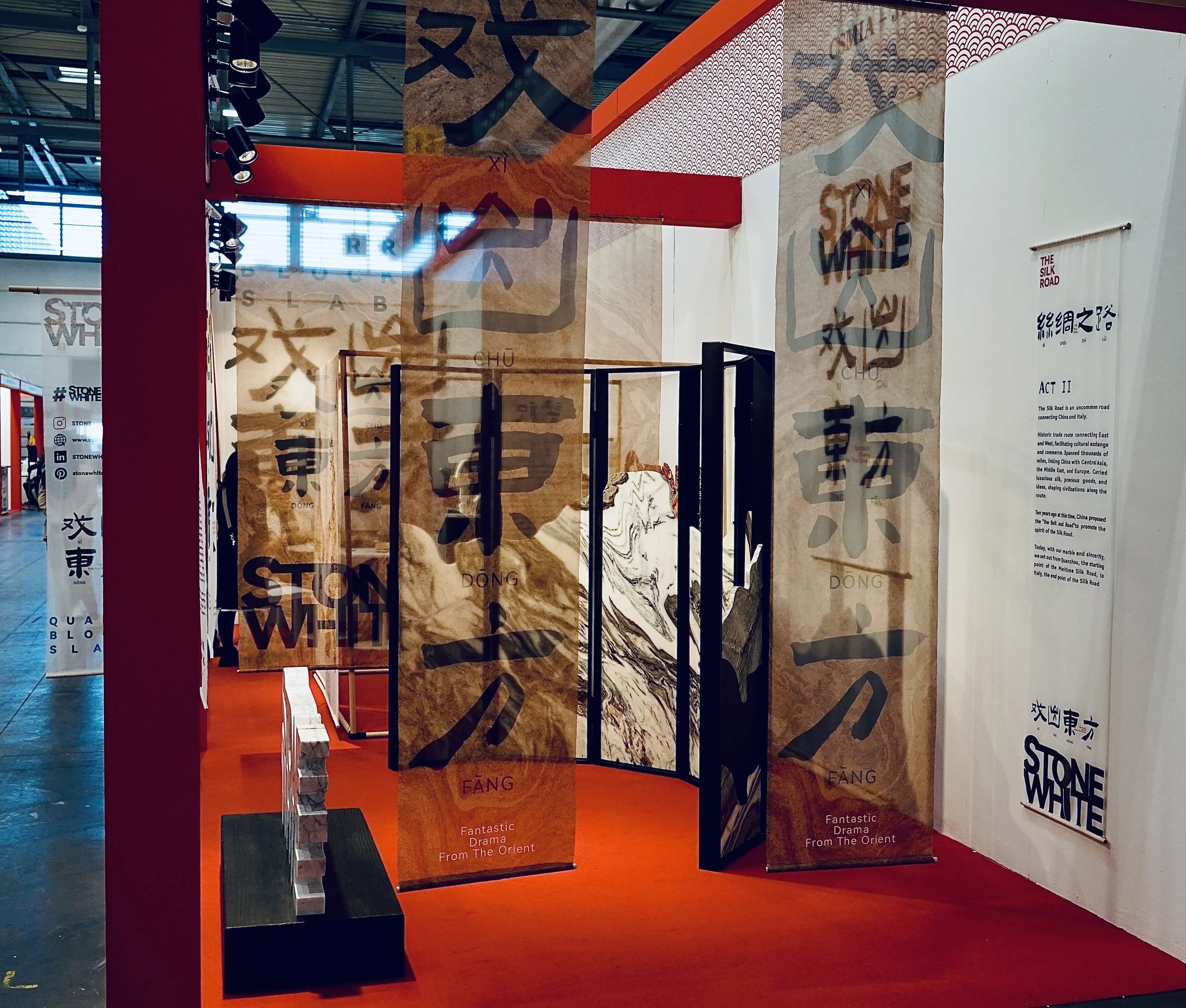 Exhibition display with large banners featuring Chinese characters and English text 'Stone White' and 'Fantastic Drama From The Orient,' set against a red floor and white walls.