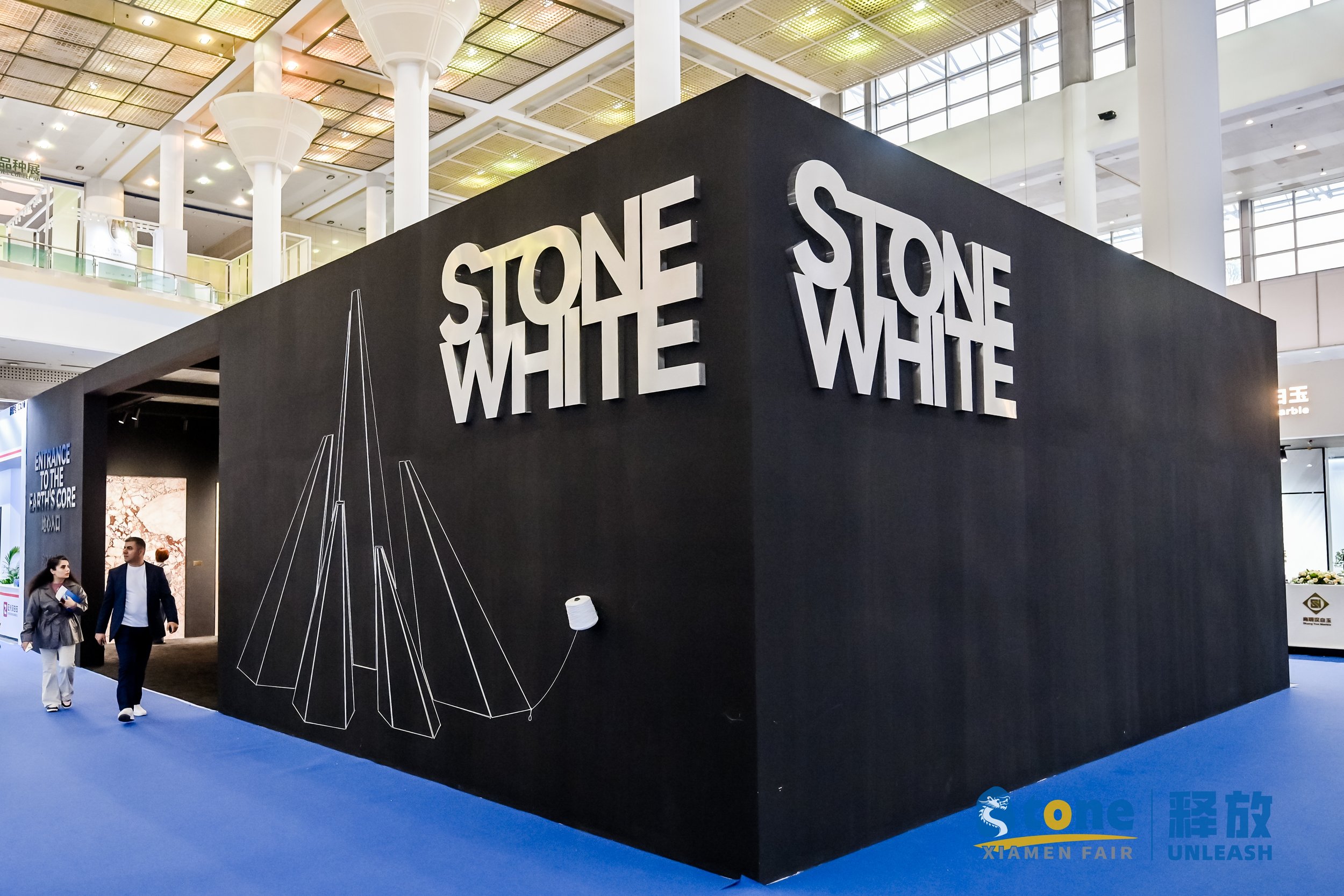 Large black exhibition booth with white 3D letters spelling 'STONE WHITE' on the walls, inside a spacious, modern indoor venue with high ceilings and glass windows. Two people are walking near the booth, and a colorful graphics setup is visible in th