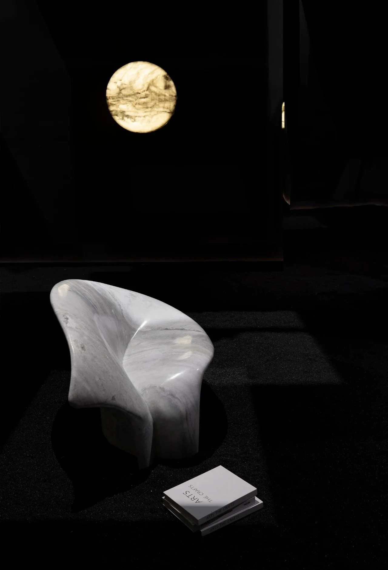 A modern, abstract white marble chair with a smooth, curved design sits on black flooring. In front of it, there are small, stacked booklets or cards, one of which reads 'ARTS THE CRAFTS.' In the background, a round, illuminated artwork or window fea