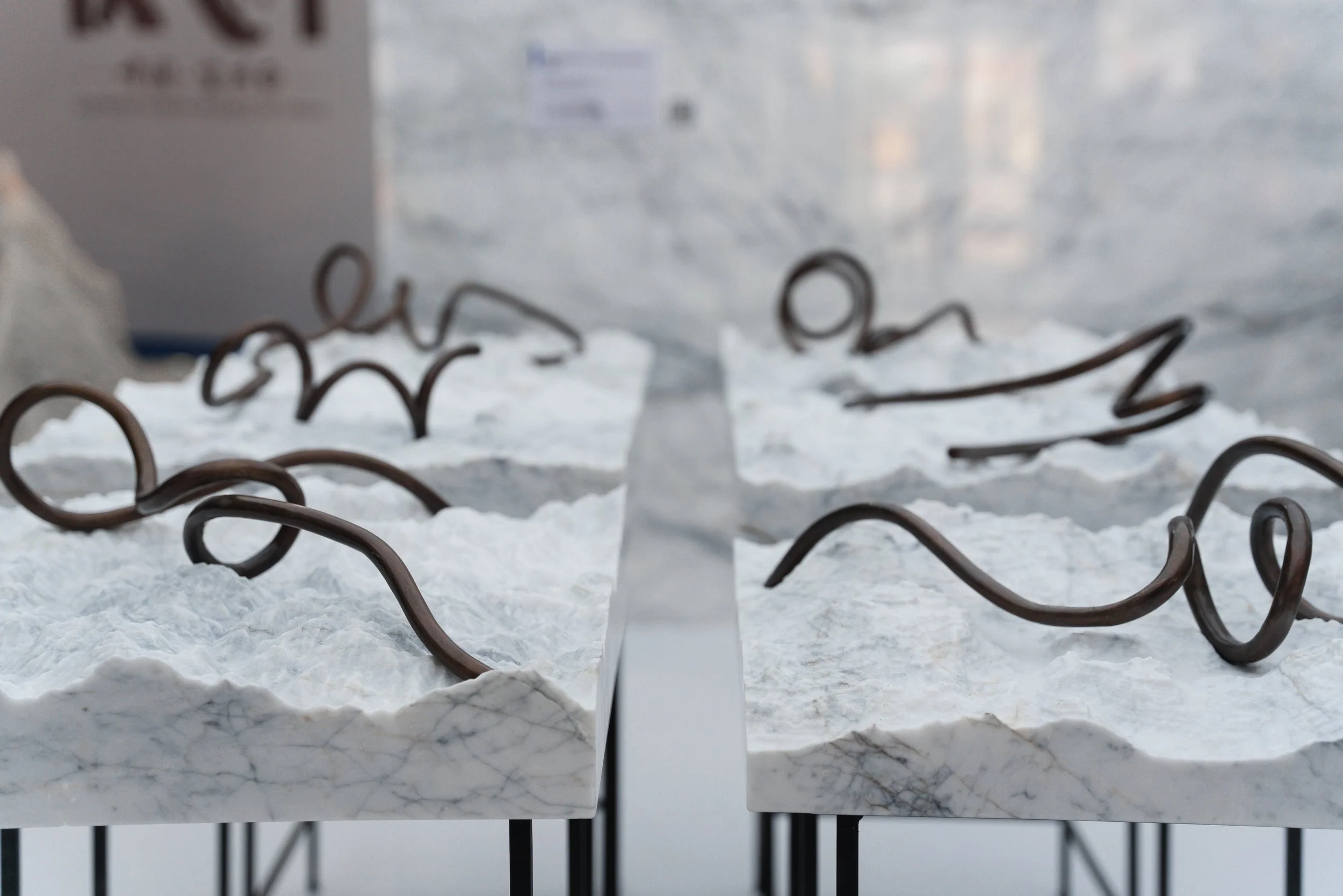 Close-up of a marble surface with dark metal wires arranged in a wavy pattern on top, reflected in a mirror showing a slightly blurred background with boxes.