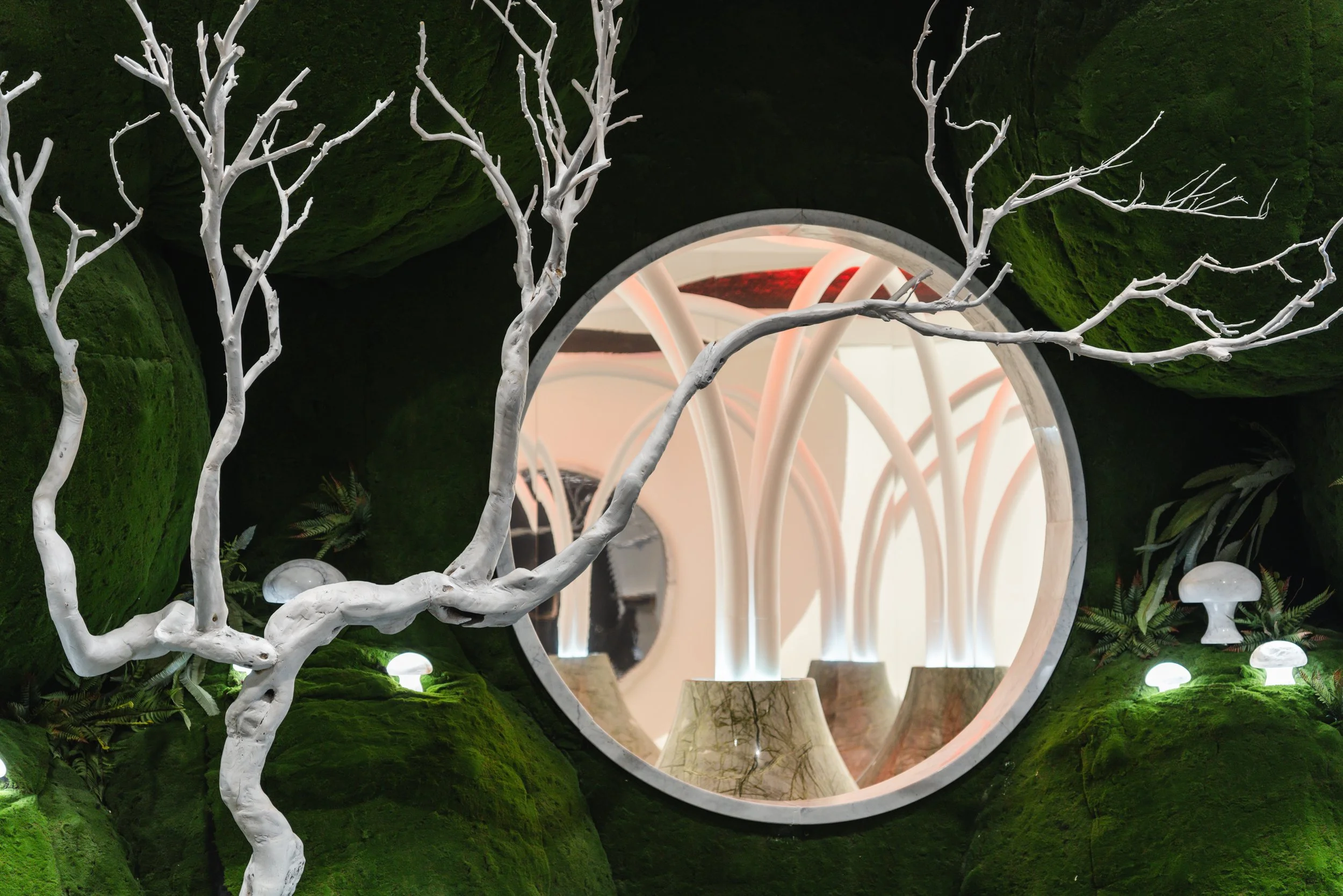 An interior scene featuring a moss-covered green wall with white, leafless tree branches extending through a circular window, which is reflecting a modern space with flowing, curved white structures and small mushroom-like decorations.